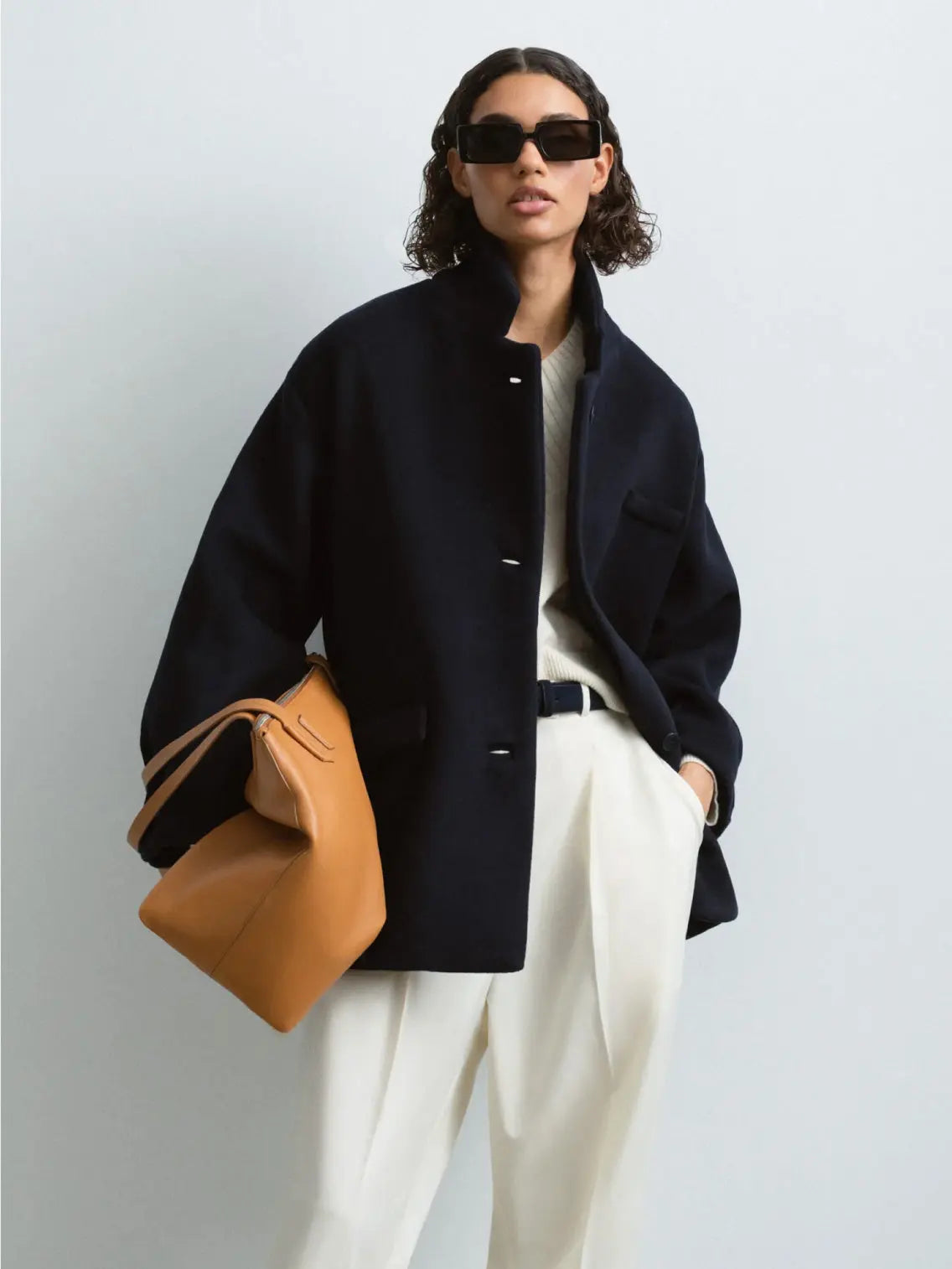 A person with shoulder-length curly hair wears dark sunglasses, a Wool Coat Navy by Cordera, and white pants. They hold a large tan handbag in one hand, exuding a relaxed, confident stance as if they just stepped out of a trendy Barcelona store against a plain background.