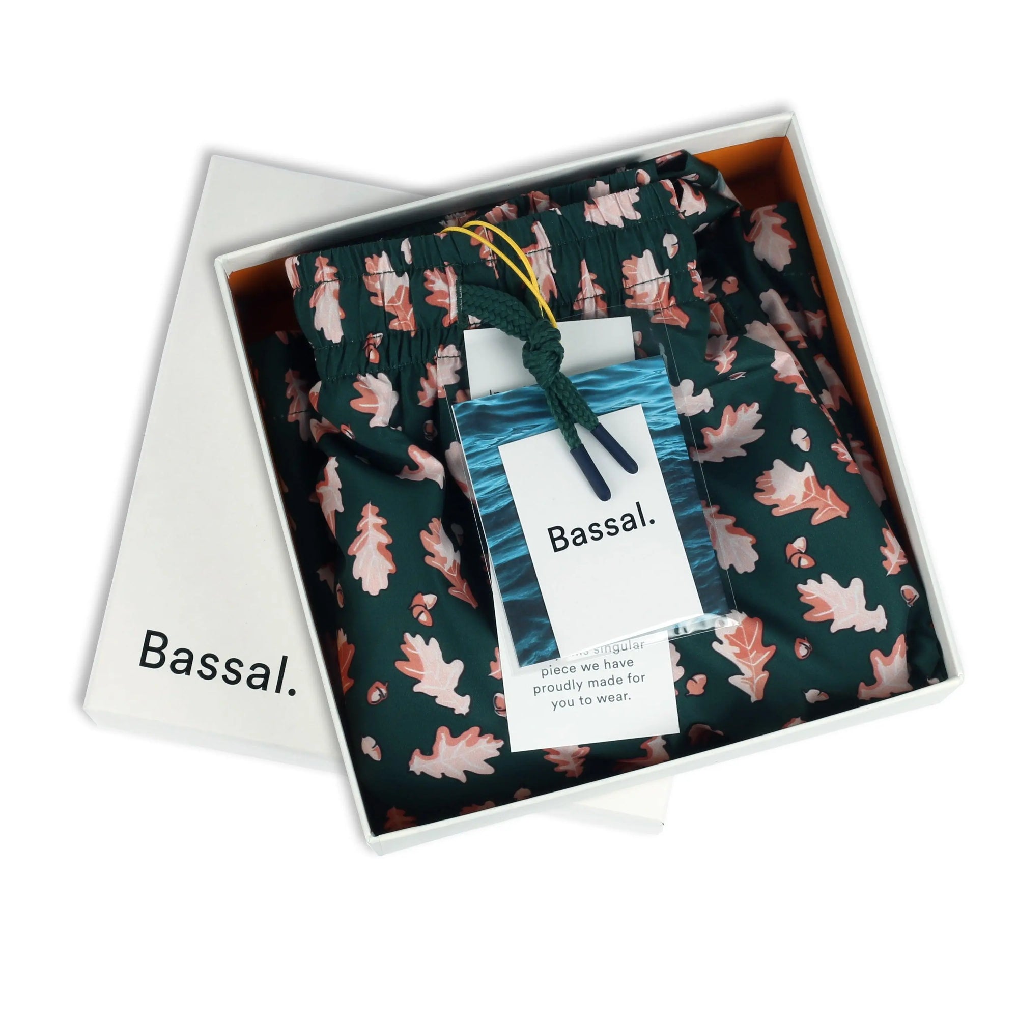 An open white box displays a pair of folded navy blue Acorn Swimwear adorned with a pink leaf pattern. Attached to the swimwear is a white tag bearing the brand name "Bassal." The partially visible box lid in the background also reads "Bassal." This item is available exclusively at bassalstore in Barcelona.