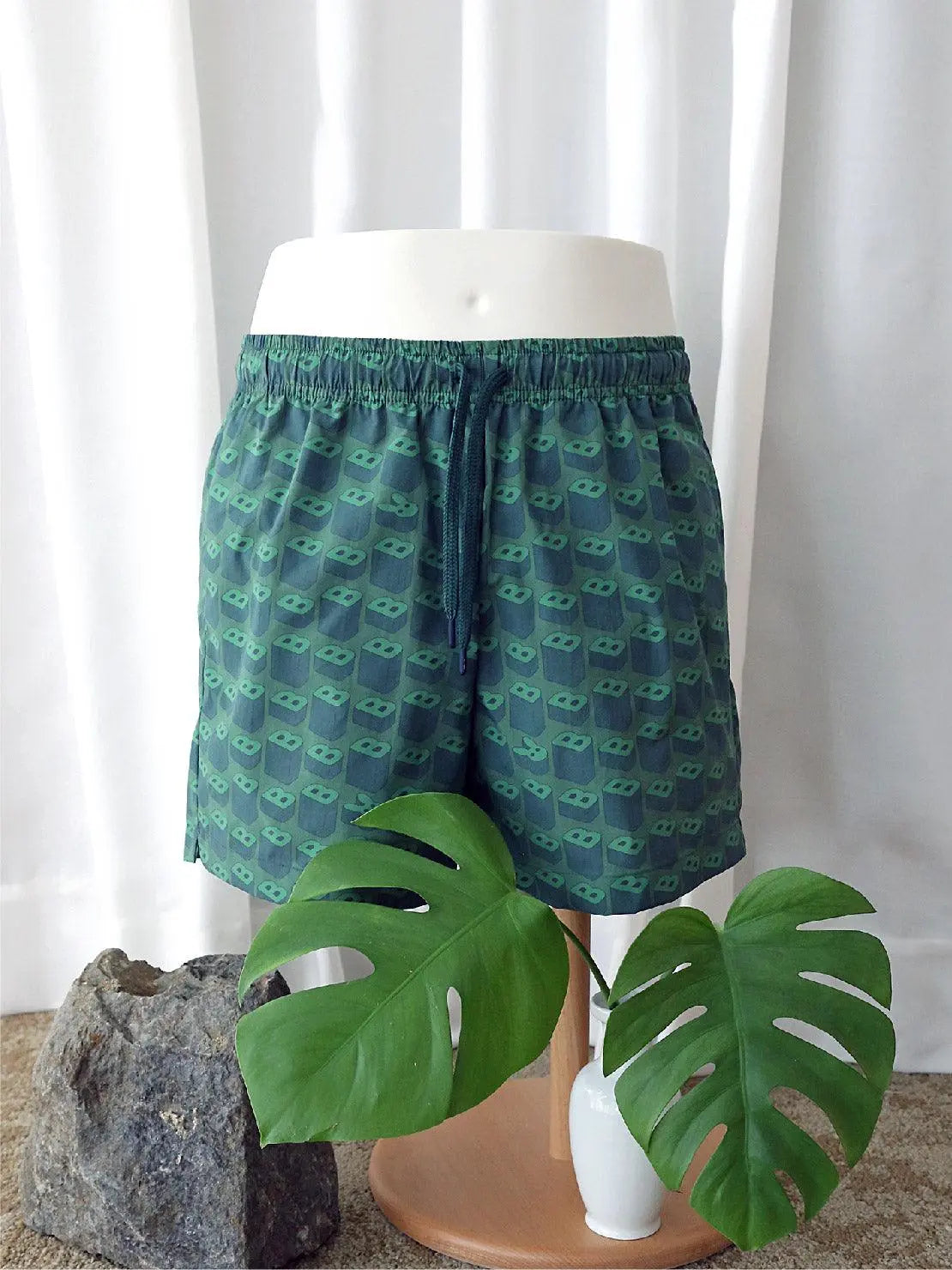 A pair of B's Green Swimwear with a pattern of small green 3D glasses displayed on a mannequin against a white curtain backdrop. The swimwear has an elastic waistband with a drawstring. In the foreground, there is a large green monstera leaf and a small rock on a circular wooden stand from Bassal in Barcelona.