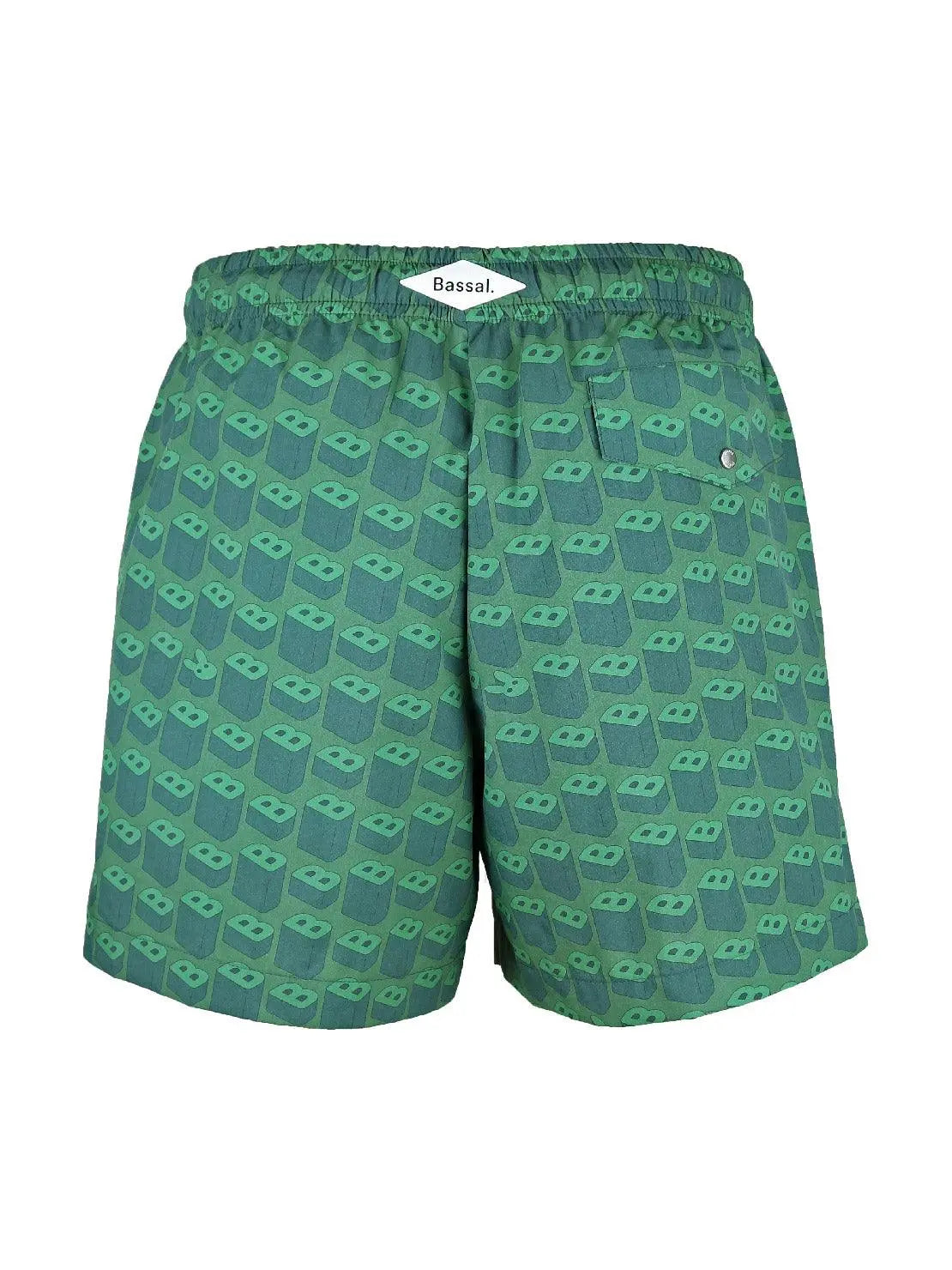 A pair of B's Green Swimwear with a pattern of small green 3D glasses displayed on a mannequin against a white curtain backdrop. The swimwear has an elastic waistband with a drawstring. In the foreground, there is a large green monstera leaf and a small rock on a circular wooden stand from Bassal in Barcelona.