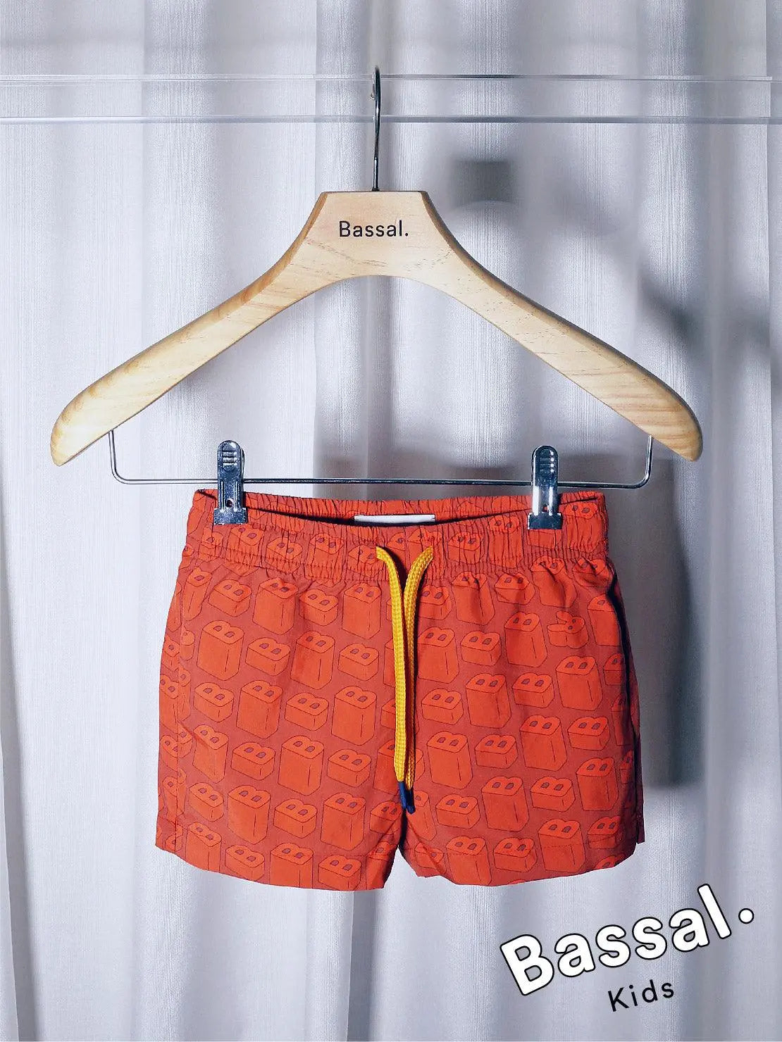 Bright orange children's swim shorts with a happy cartoon face pattern hang on a wooden hanger labeled "Bassal." The shorts, known as B's Red Kids Swimwear, feature a yellow drawstring and metallic clips, with the brand name "Bassal" displayed in the bottom right corner. Available at bassalstore in Barcelona.