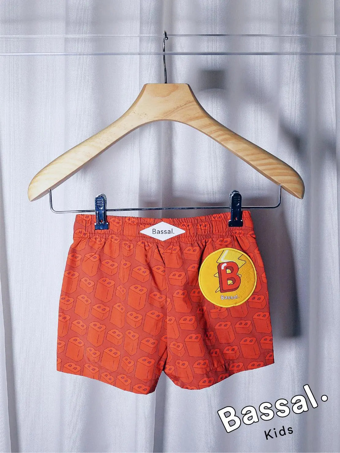 Bright orange children's swim shorts with a happy cartoon face pattern hang on a wooden hanger labeled "Bassal." The shorts, known as B's Red Kids Swimwear, feature a yellow drawstring and metallic clips, with the brand name "Bassal" displayed in the bottom right corner. Available at bassalstore in Barcelona.