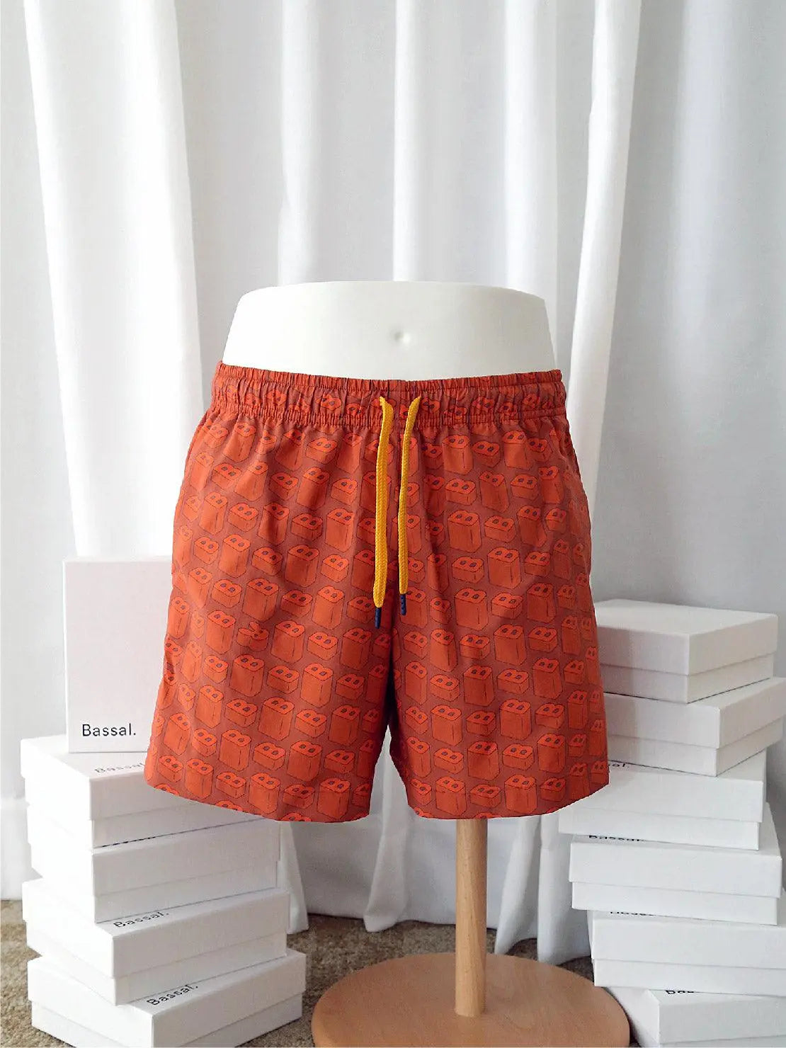 A pair of B's Red Swimwear with a pattern of small teddy bears displayed on a mannequin. The swimwear has a yellow drawstring at the waist. The background features a white curtain and several neatly stacked white boxes, one of which displays the brand name "Bassal" from the famed bassalstore in Barcelona.