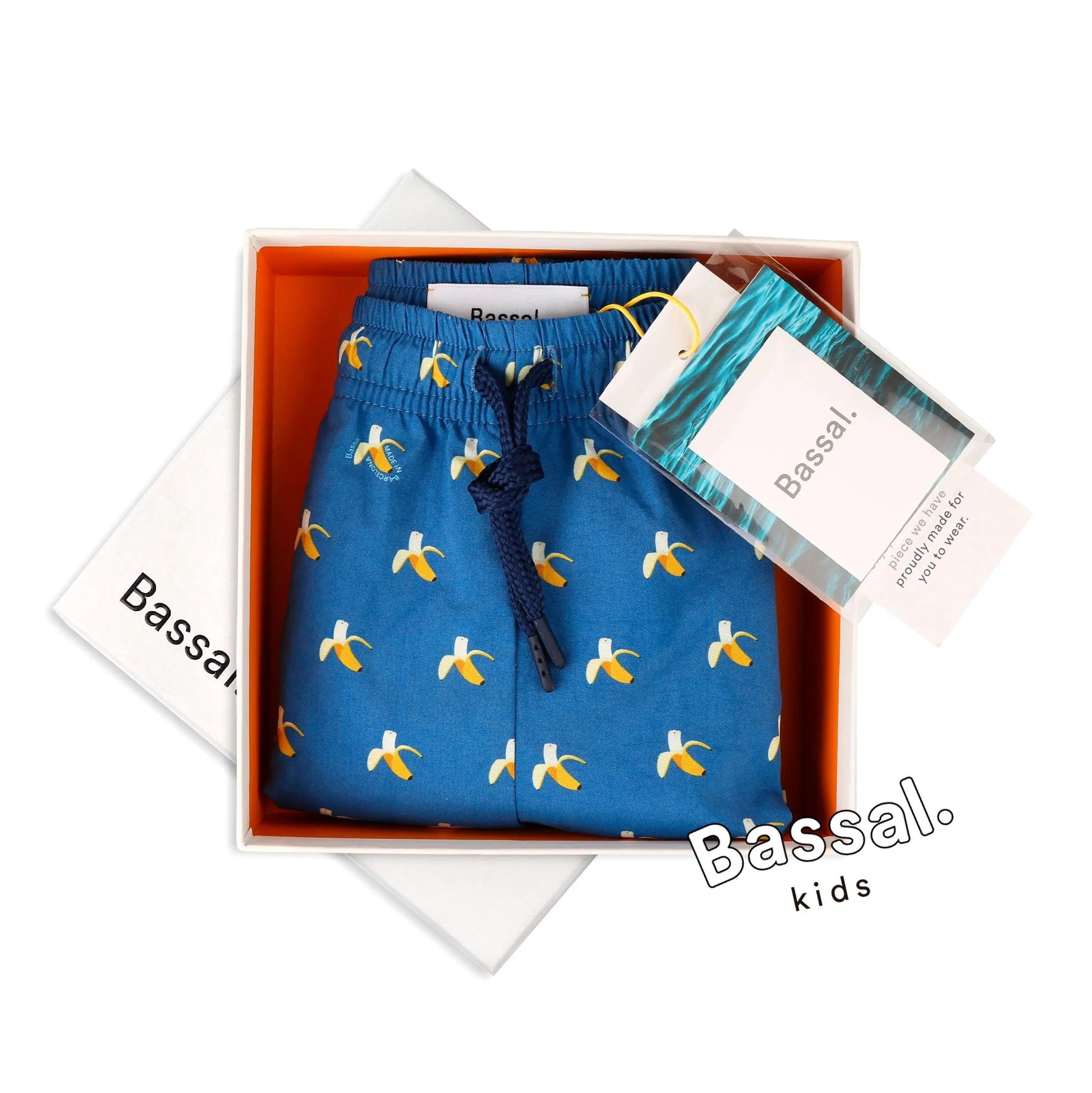 A pair of blue Bananas Kids Swimwear with a yellow and white banana print is neatly displayed in an orange-lined gift box. The swimwear has a navy blue drawstring and is tagged with a Bassal. kids label. The box lid features the brand name "Bassal" from bassalstore, your go-to store for fun fashion.
