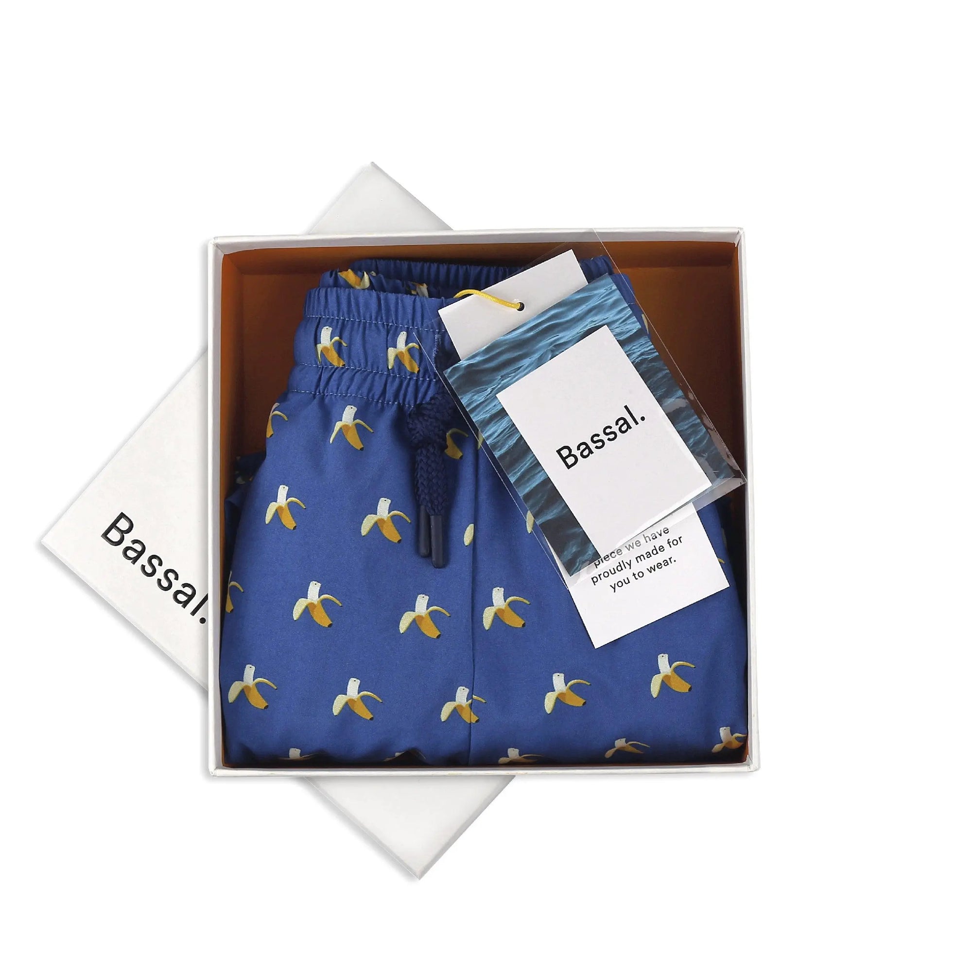 A pair of blue shorts with a banana pattern, neatly folded in a white box. The box lid, partially visible, has "Bassal." written on it. Also inside the box are two labeled packets, one possibly containing a product tag and the other in blue packaging. This delightful item, Bananas Swimwear by Bassal., is available at Bassalstore in Barcelona.