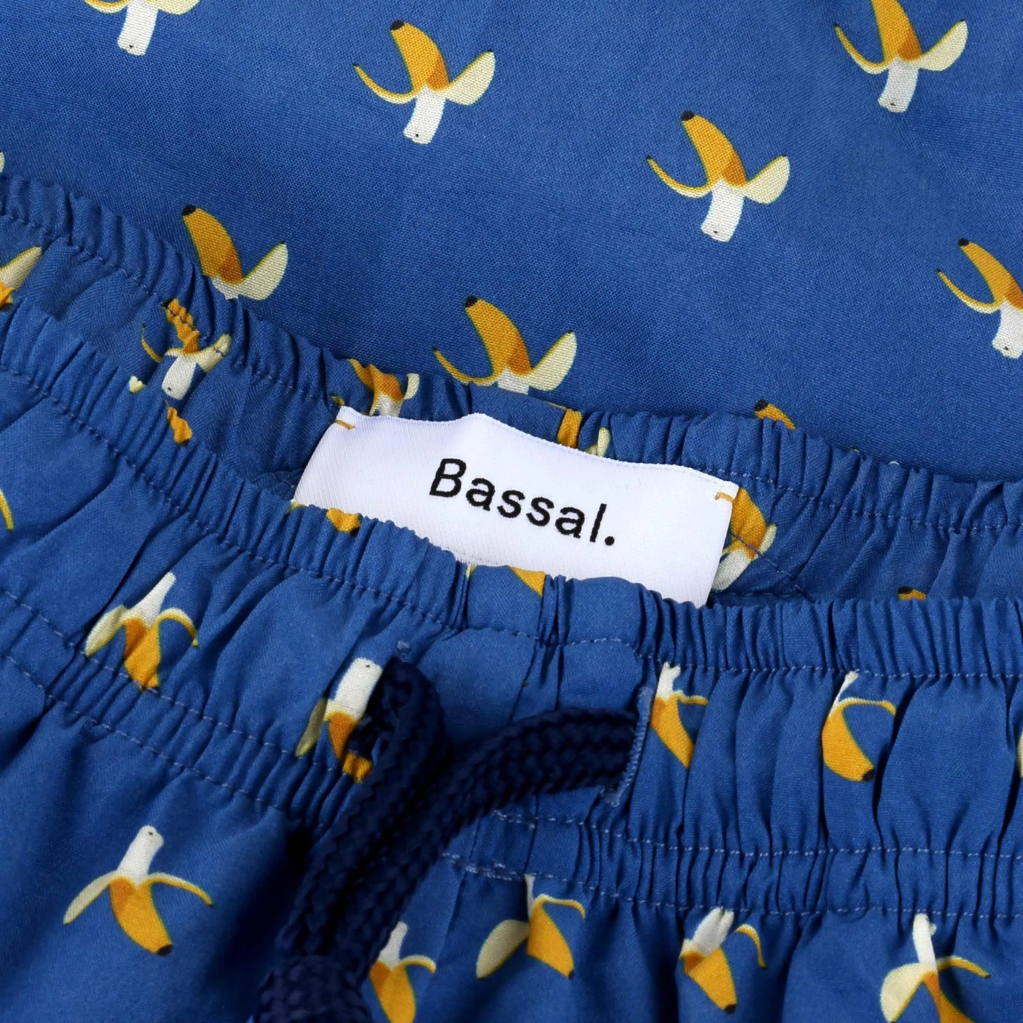 A pair of blue shorts with a banana pattern, neatly folded in a white box. The box lid, partially visible, has "Bassal." written on it. Also inside the box are two labeled packets, one possibly containing a product tag and the other in blue packaging. This delightful item, Bananas Swimwear by Bassal., is available at Bassalstore in Barcelona.