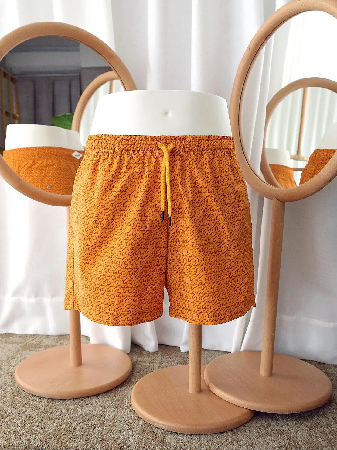 A mannequin at Bassalstore in Barcelona wears Bassal Orange Pattern Swimwear from the brand Bassal. Positioned between two round mirrors on wooden stands, the mannequin showcases reflections of the swimwear from multiple angles. A white curtain hangs in the background.