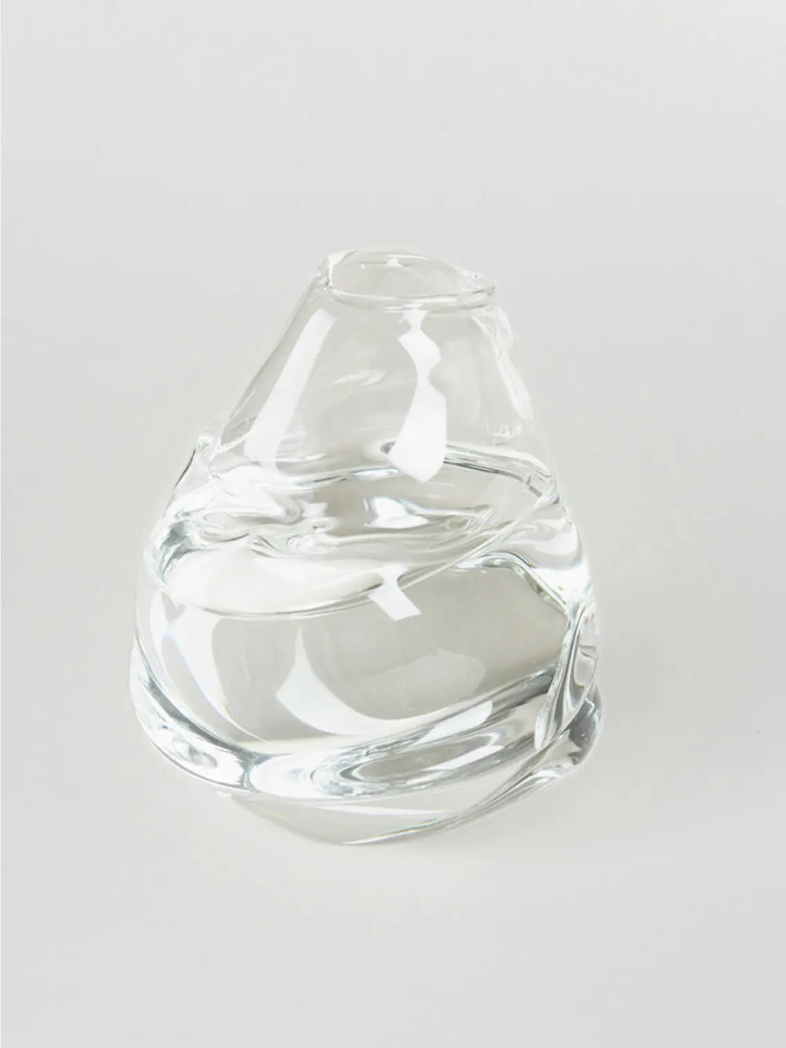 A small, clear glass vase with a slightly tilted, asymmetrical shape is shown against a plain white background. The vase has a smooth, modern design and is empty inside—a perfect piece you might find at Bassalstore in Barcelona. This is the Ben Jor Vase Clear by Nathalie Schreckenberg.