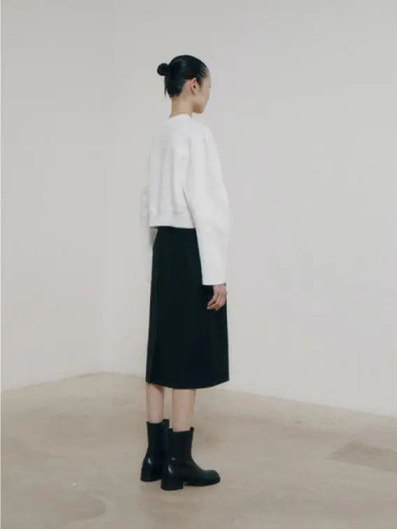 A person with tied-back hair stands indoors against a plain backdrop, wearing a loose-fitting white long-sleeved top from Bassalstore, an Amomento Black Volume Skirt, and black ankle boots. They are facing forward, with a neutral expression and their arms relaxed by their sides.