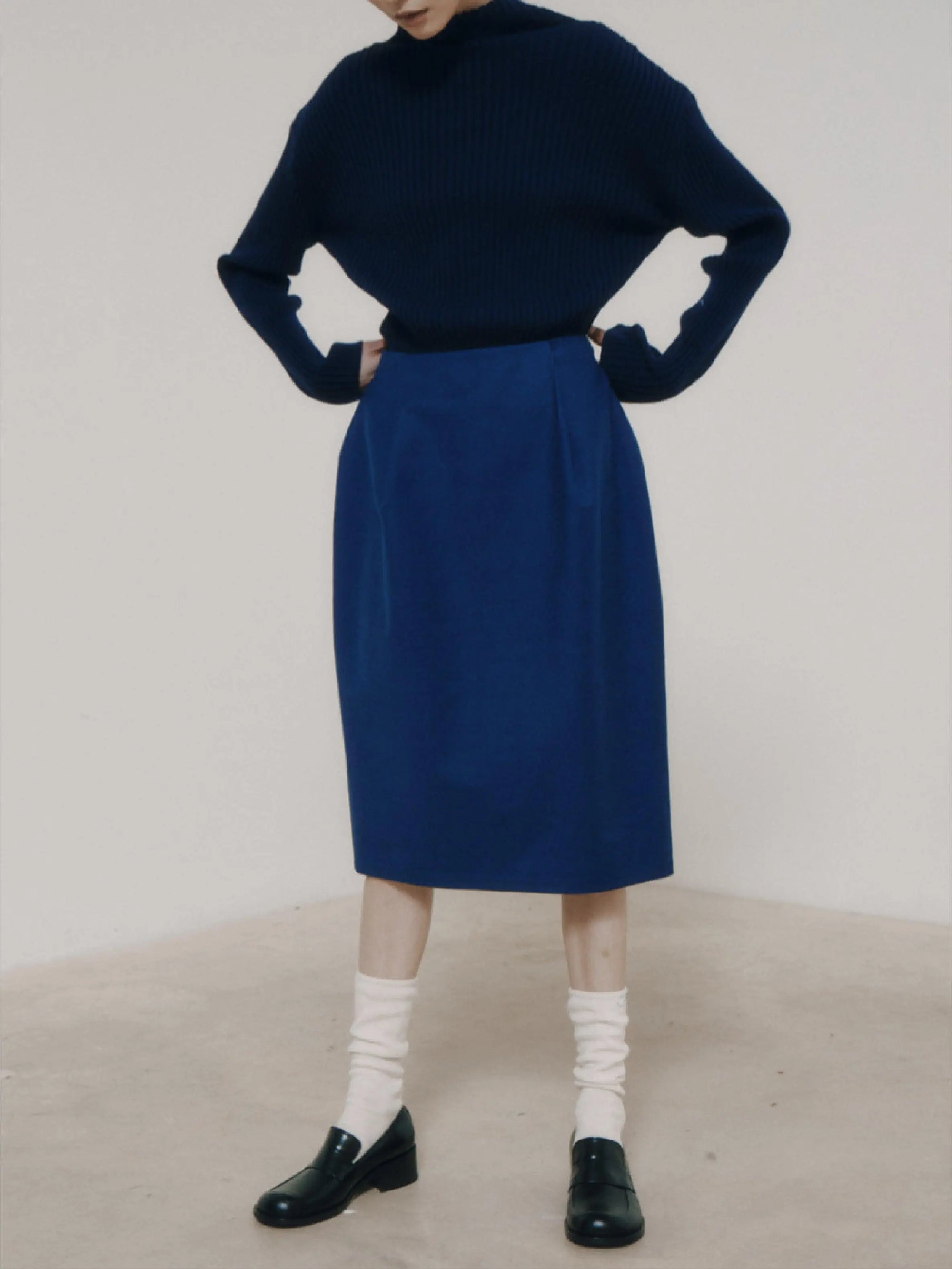A person wearing a dark blue long-sleeved top is paired with the Amomento Blue Volume Skirt. The outfit, from bassalstore, is complemented by white socks and black loafers. The individual has their hands on their hips and is standing against a neutral background in Barcelona. The face is out of the frame.