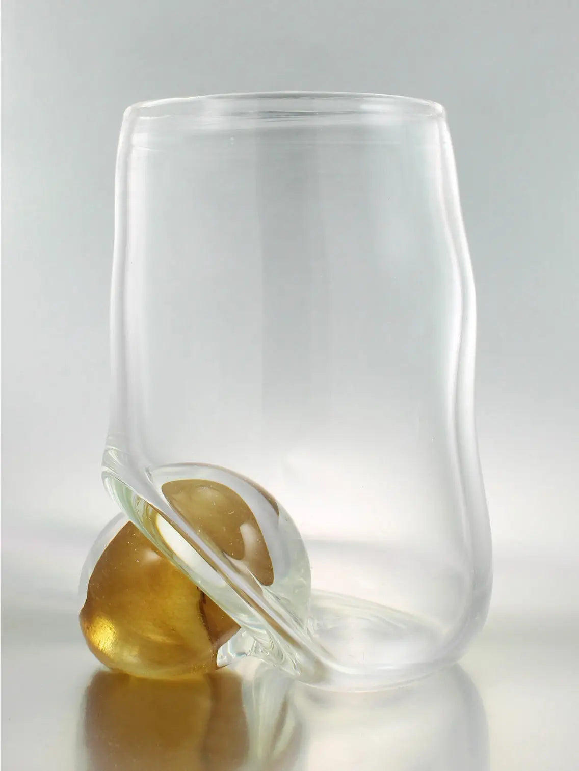 A Bola Vase from Nathalie Schreckenberg in Barcelona, featuring a clear, wavy cylindrical form with a rounded base. Encased within it is a spherical object with amber coloration, creating a unique blend of transparency and color. The setting is a simple, light background.