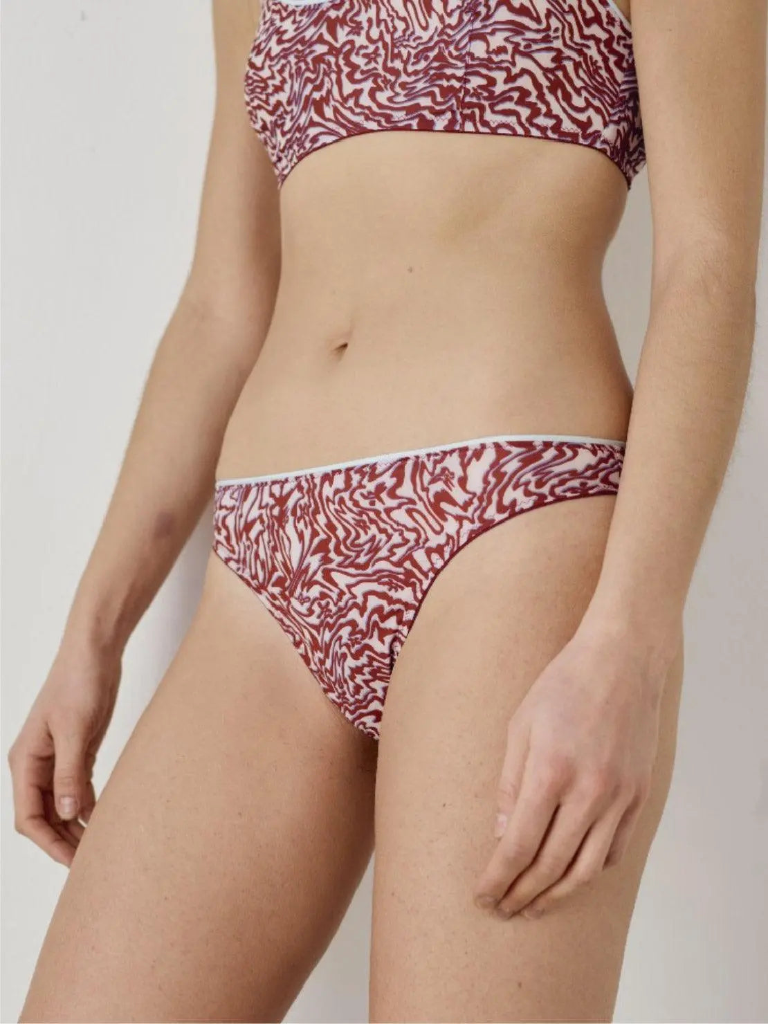 A person is wearing a matching set of red and white patterned underwear from Interiors, consisting of a bralette and a pair of Brazilian Briefs. The image shows the person's torso and part of their upper thighs. The background is plain and uncluttered.