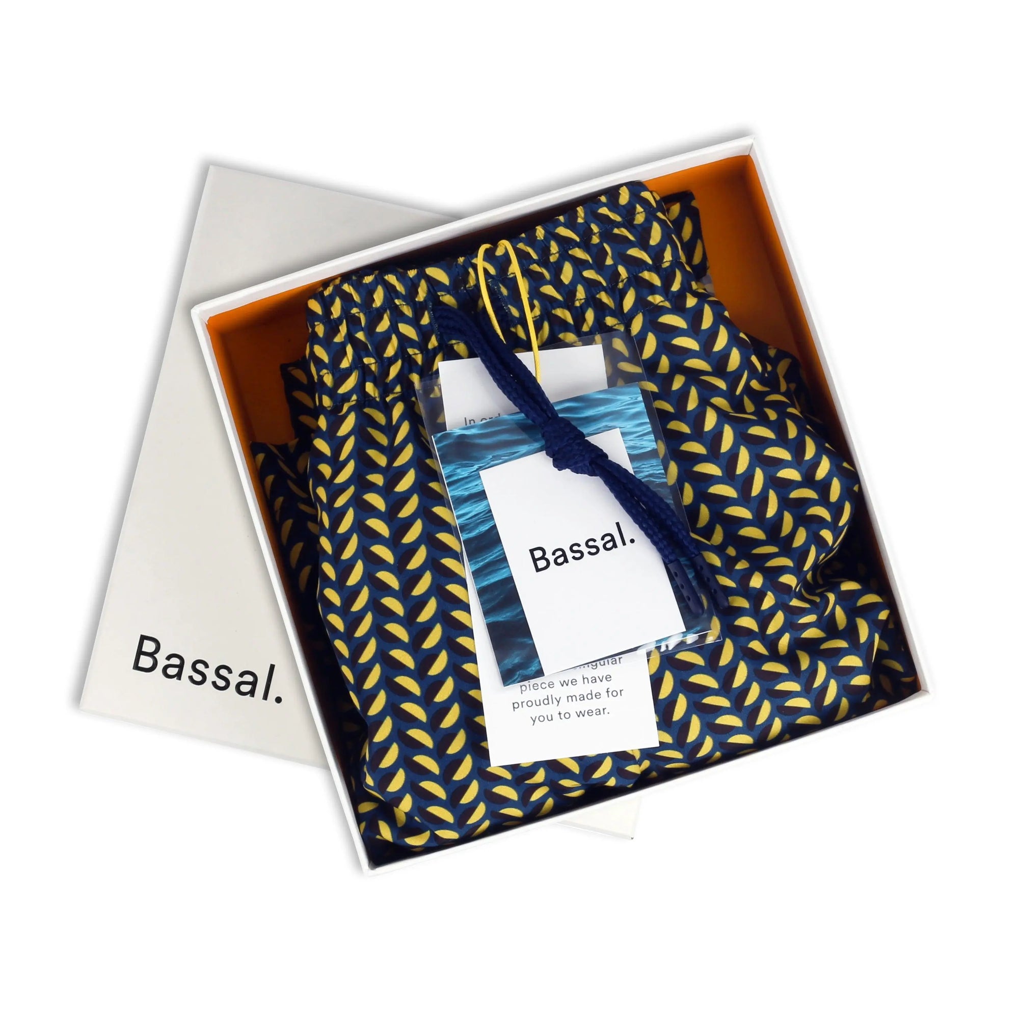 A neatly packaged pair of Clams Blue Swimwear with a blue and yellow leaf pattern, partially inside an open white box. The box lid, placed askew, displays the brand name "Bassal." A small tag with "bassalstore" branding is attached to the shorts with a yellow drawstring.