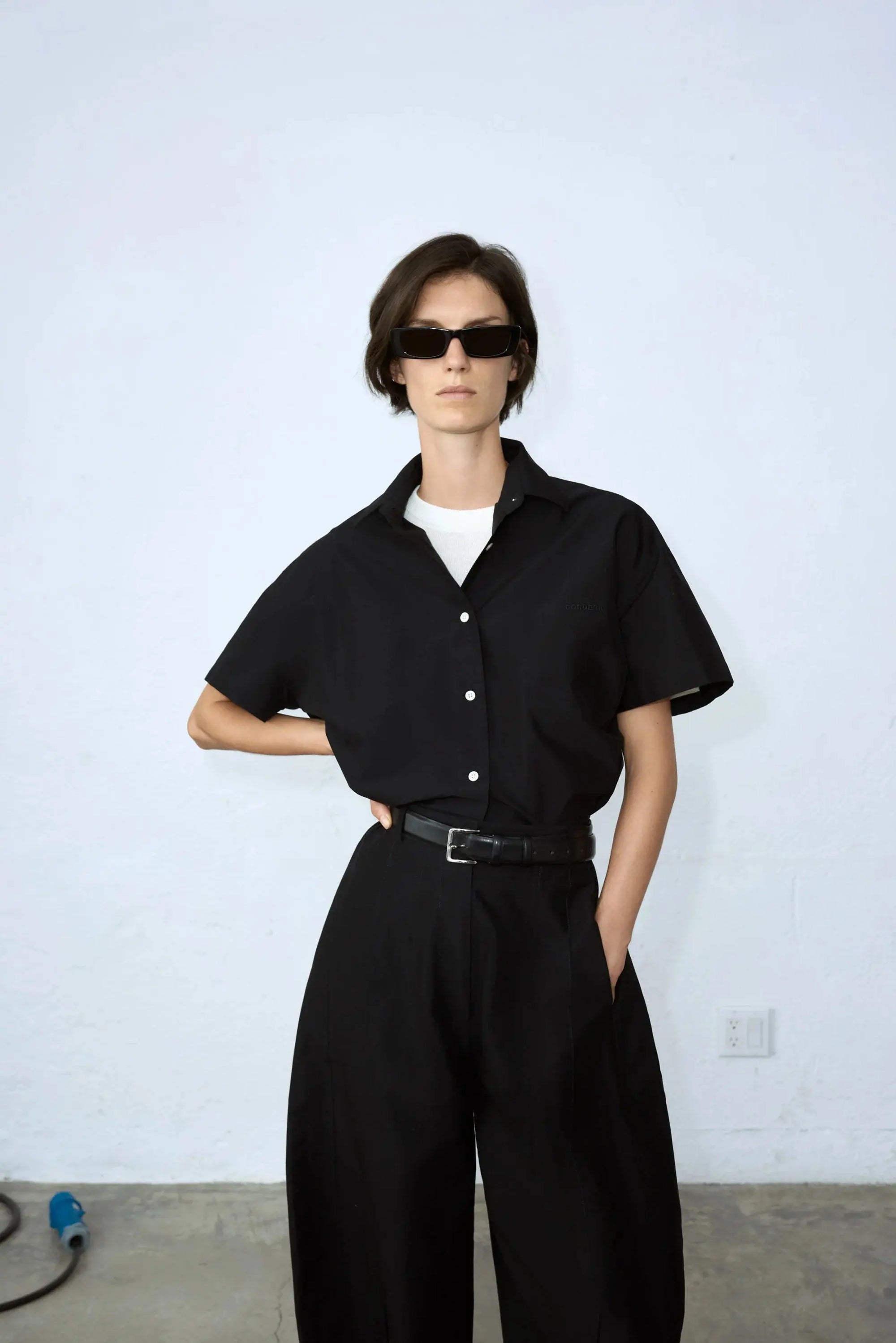 A black Cropped Shirt Black with a collar, buttoned up at the front with white buttons. It has a small front pocket and a tag inside the collar that reads "Cordera" in uppercase letters. The shirt is displayed against a plain white background and is available at bassalstore, Barcelona.
