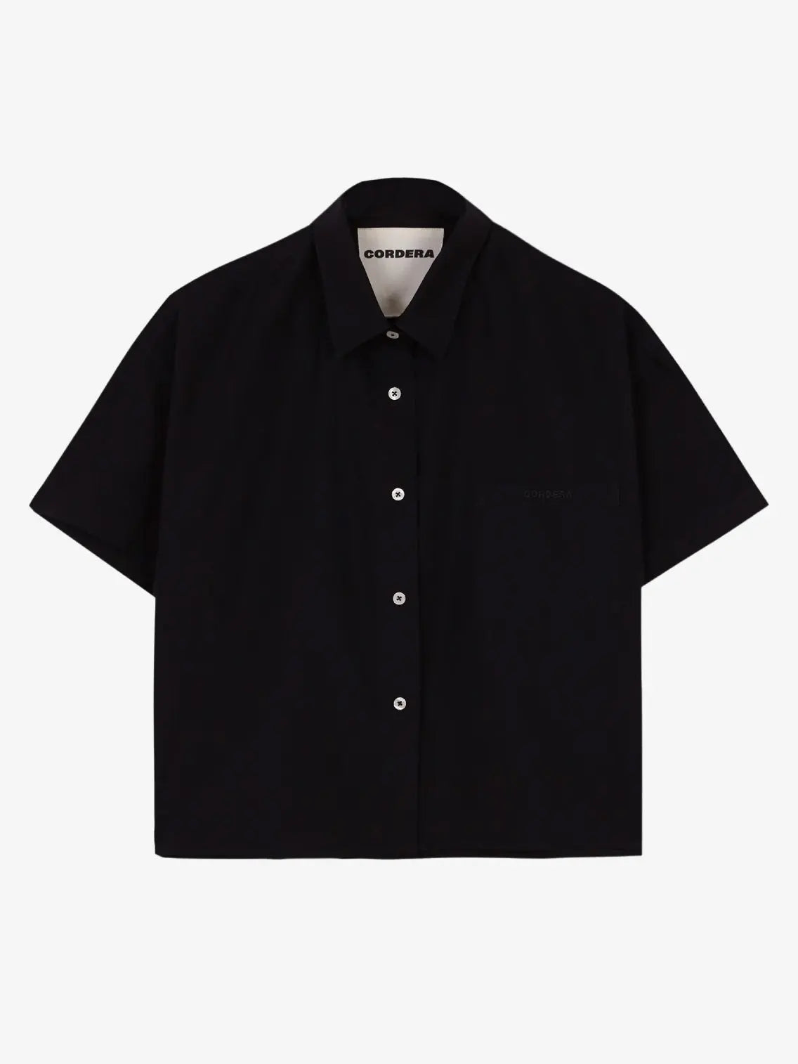 A black Cropped Shirt Black with a collar, buttoned up at the front with white buttons. It has a small front pocket and a tag inside the collar that reads "Cordera" in uppercase letters. The shirt is displayed against a plain white background and is available at bassalstore, Barcelona.