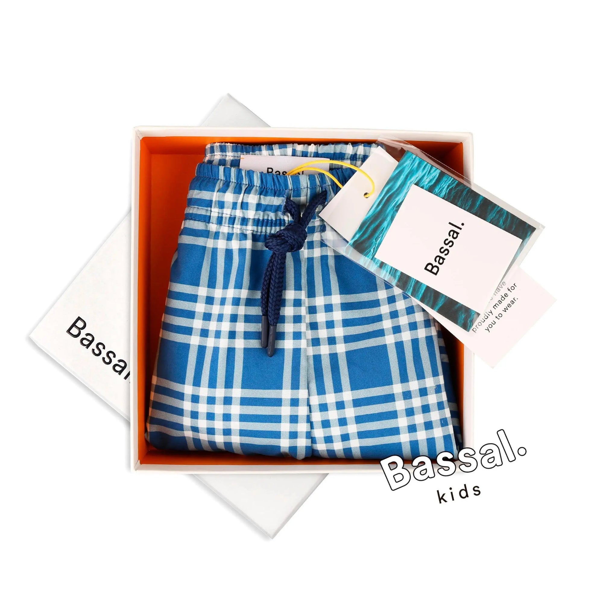 A neatly folded pair of blue and white plaid Cuatro Kids Swimwear with a navy blue drawstring is displayed inside an orange-bordered gift box. The box lid, featuring the brand name "Bassal." from the popular Bassalstore in Barcelona, is partially visible on the side. A tag with "Bassal. kids" is attached to the swimwear.