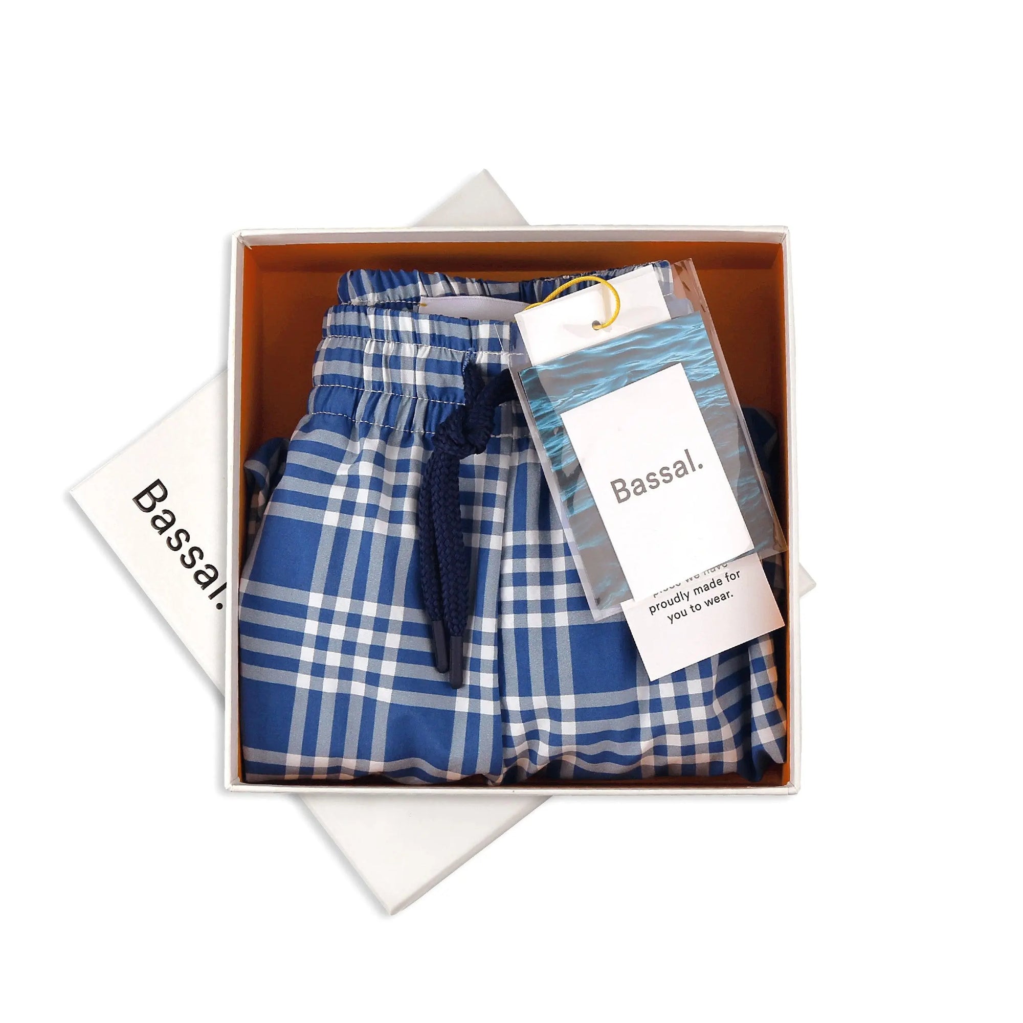 A pair of blue and white plaid Cuatro Swimwear with an elastic waistband and navy drawstring is neatly folded inside an open Bassal. box. A clear plastic tag attached to the swimwear reads "Bassal. proudly made for you to wear." Discover them at our flagship store in Barcelona or bassalstore online.