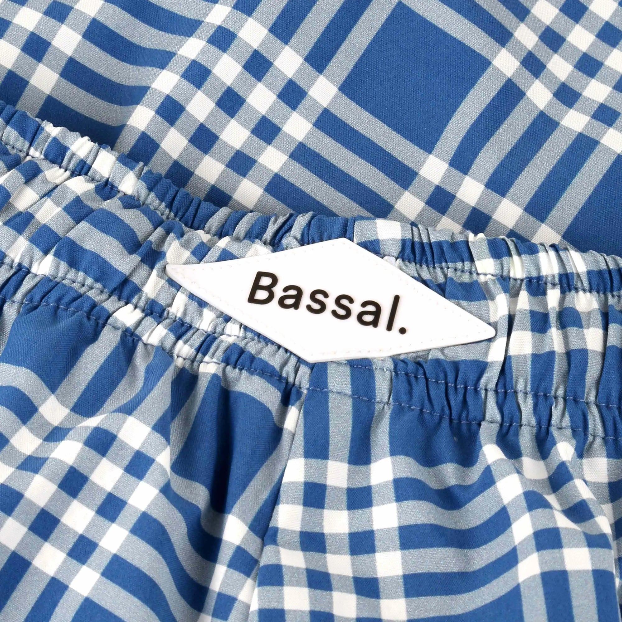 A pair of blue and white plaid Cuatro Swimwear with an elastic waistband and navy drawstring is neatly folded inside an open Bassal. box. A clear plastic tag attached to the swimwear reads "Bassal. proudly made for you to wear." Discover them at our flagship store in Barcelona or bassalstore online.