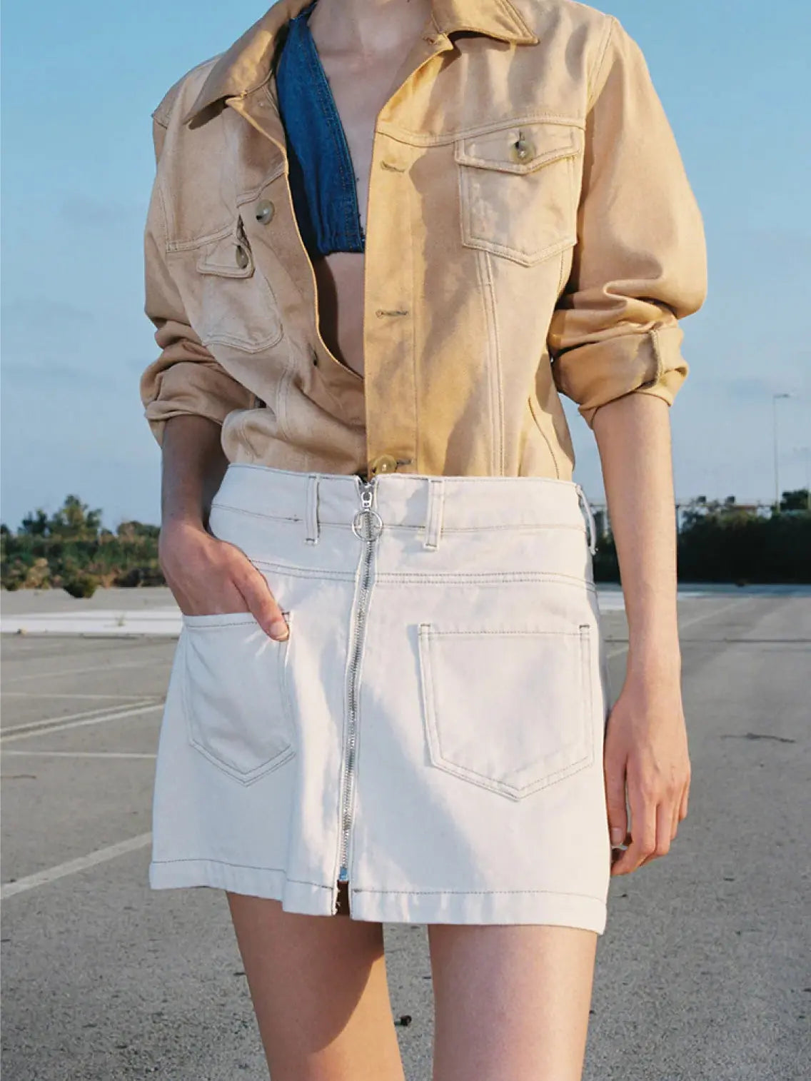 A person is standing outdoors wearing a beige buttoned-down jacket over a blue top and a Denim Mini Skirt White with front pockets and a zipper detail from Mundaka, likely bought in Bassalstore in Barcelona. The background appears to be a paved area with some greenery in the distance under a clear sky.