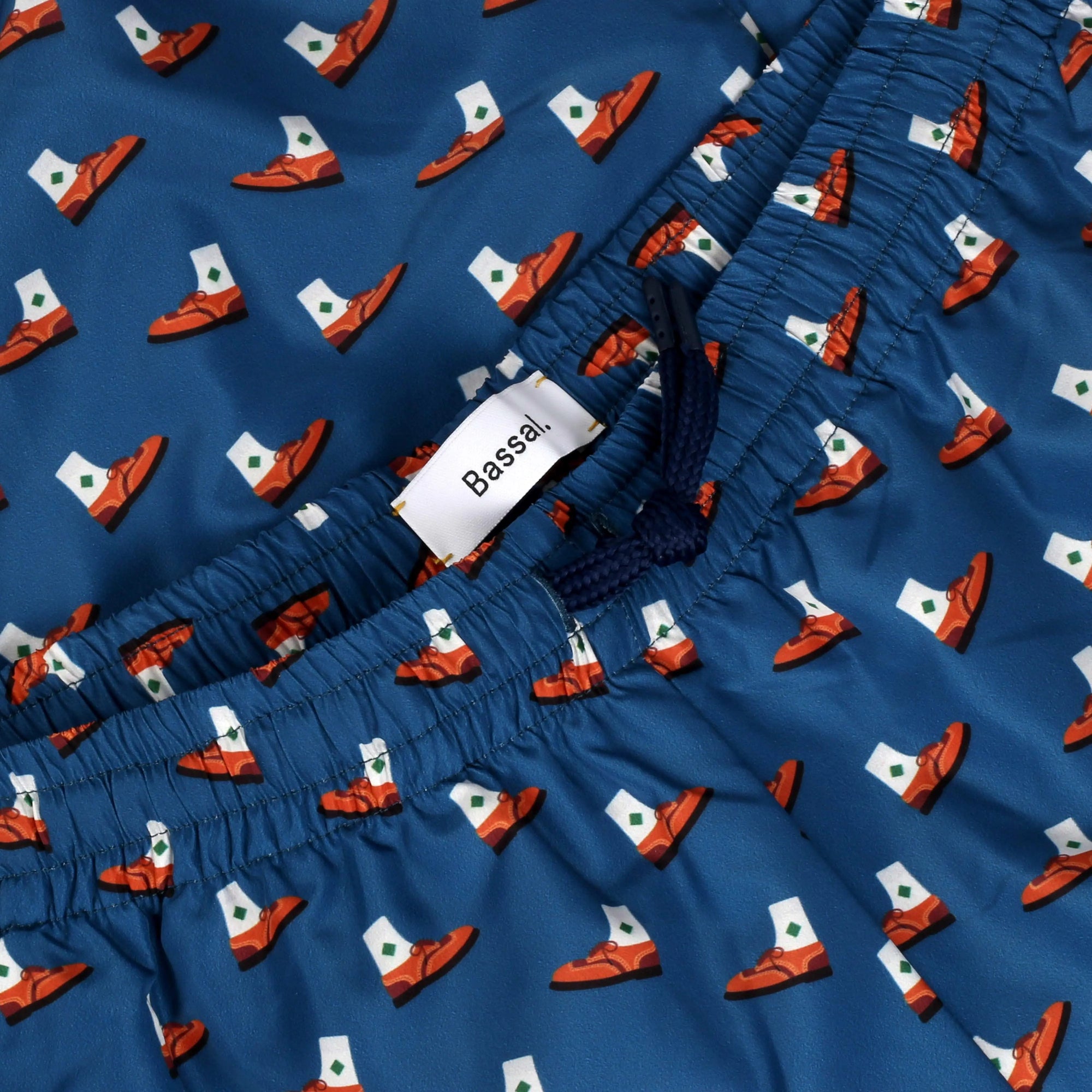 A pair of blue Derby Swimwear decorated with small boat patterns is neatly folded inside a white gift box. A clothing tag with the brand name "Bassal" hangs from the waistband. The box lid, placed to the side, also displays "Bassal." Discover more at bassalstore in Barcelona.