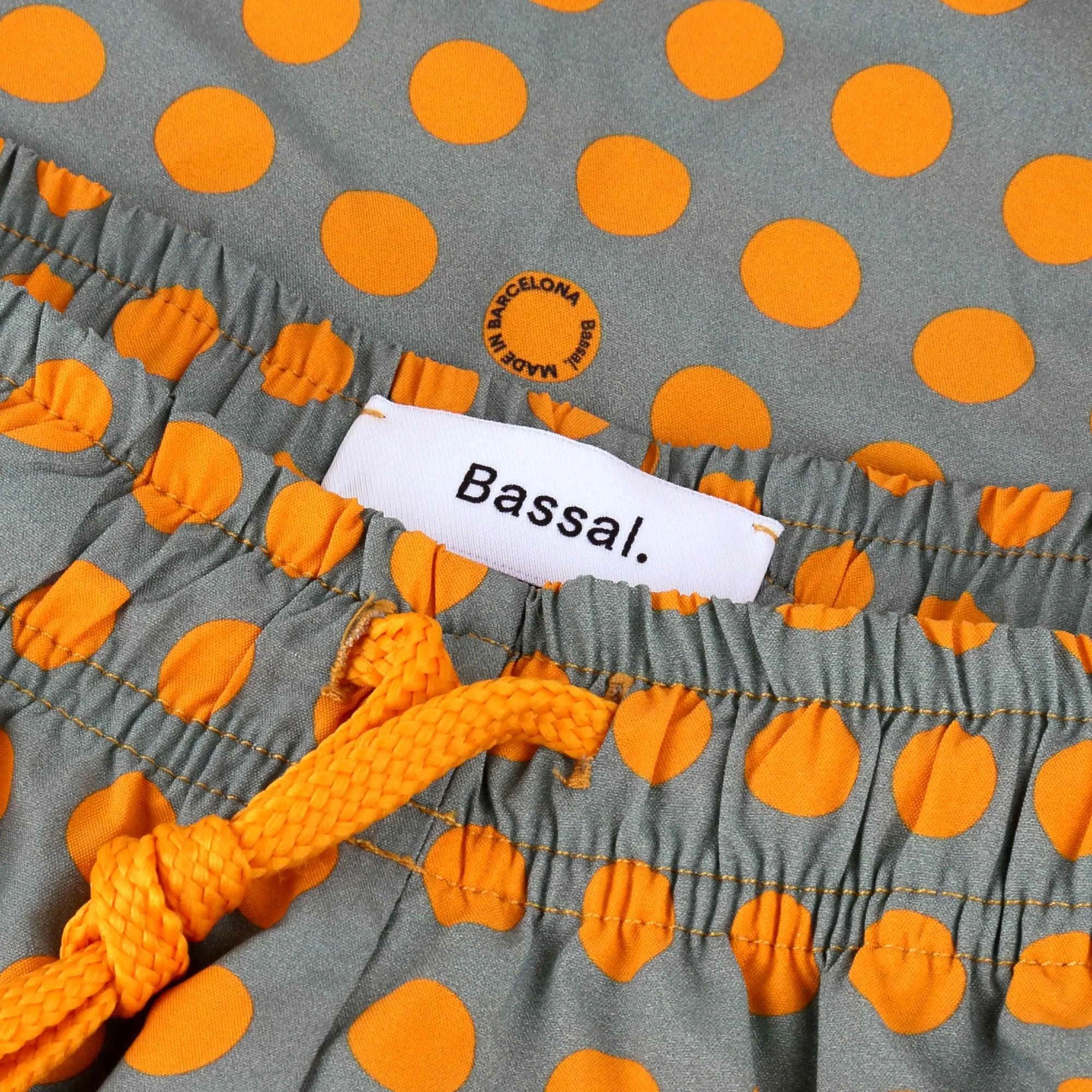 A pair of grey Dots Swimwear with orange polka dots is neatly folded in an open white box. The swimwear has an elastic waistband with a yellow drawstring. Two tags attached read "Bassal" and a note says "specifically made for you to wear." The box lid is partially visible.