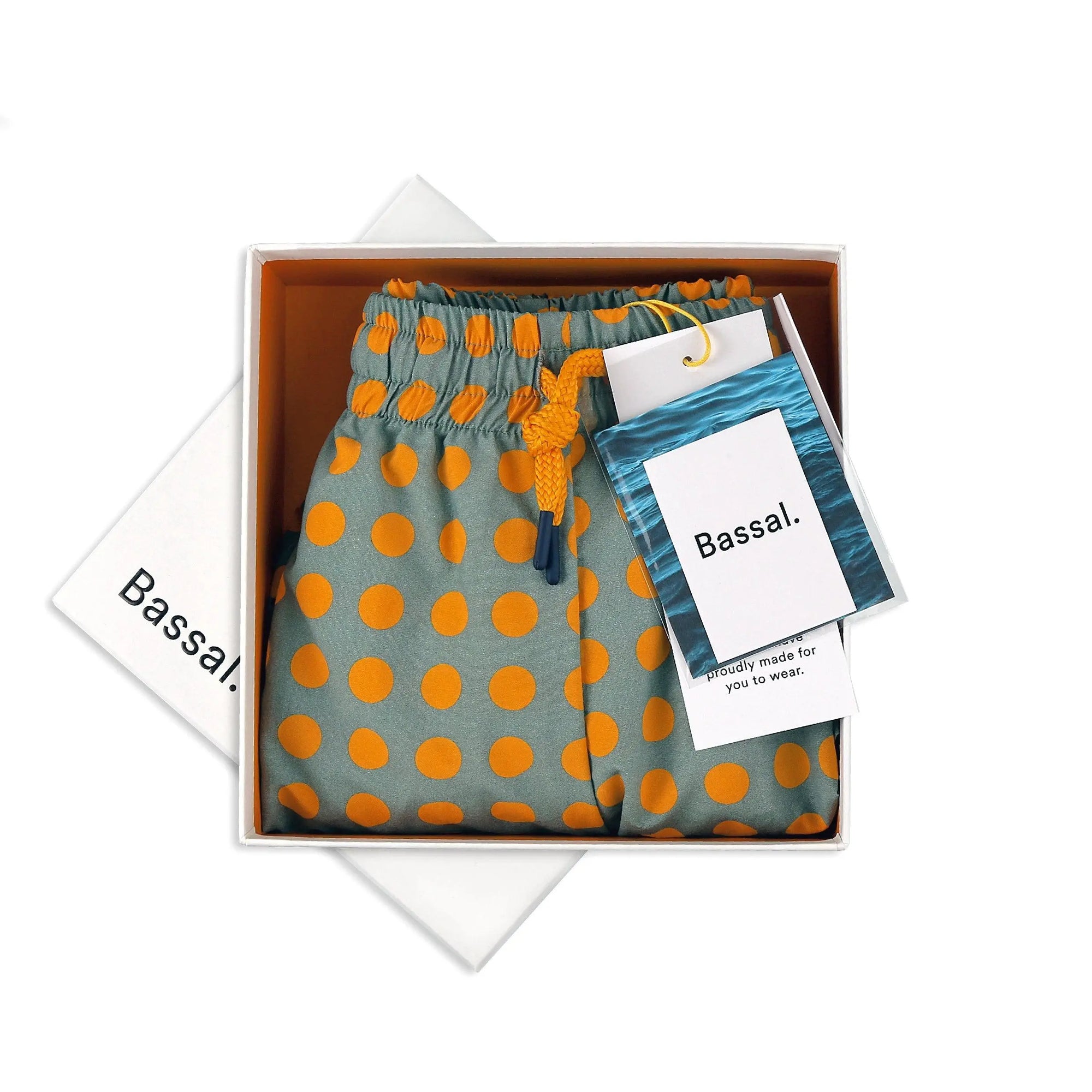 A pair of grey Dots Swimwear with orange polka dots is neatly folded in an open white box. The swimwear has an elastic waistband with a yellow drawstring. Two tags attached read "Bassal" and a note says "specifically made for you to wear." The box lid is partially visible.