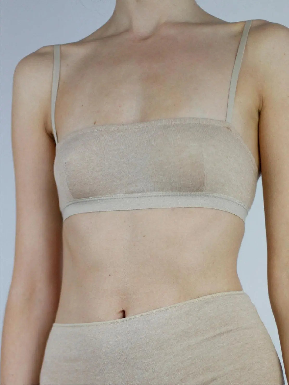 A person is shown from the shoulders to the midriff, wearing a light beige, strappy Earth Bandeau Organic Cotton Bra - Talk Under Light and high-waisted underwear. The plain gray background highlights the minimalist design of the soft, stretchable fabric. Discover this chic collection at Bassalstore in Barcelona.