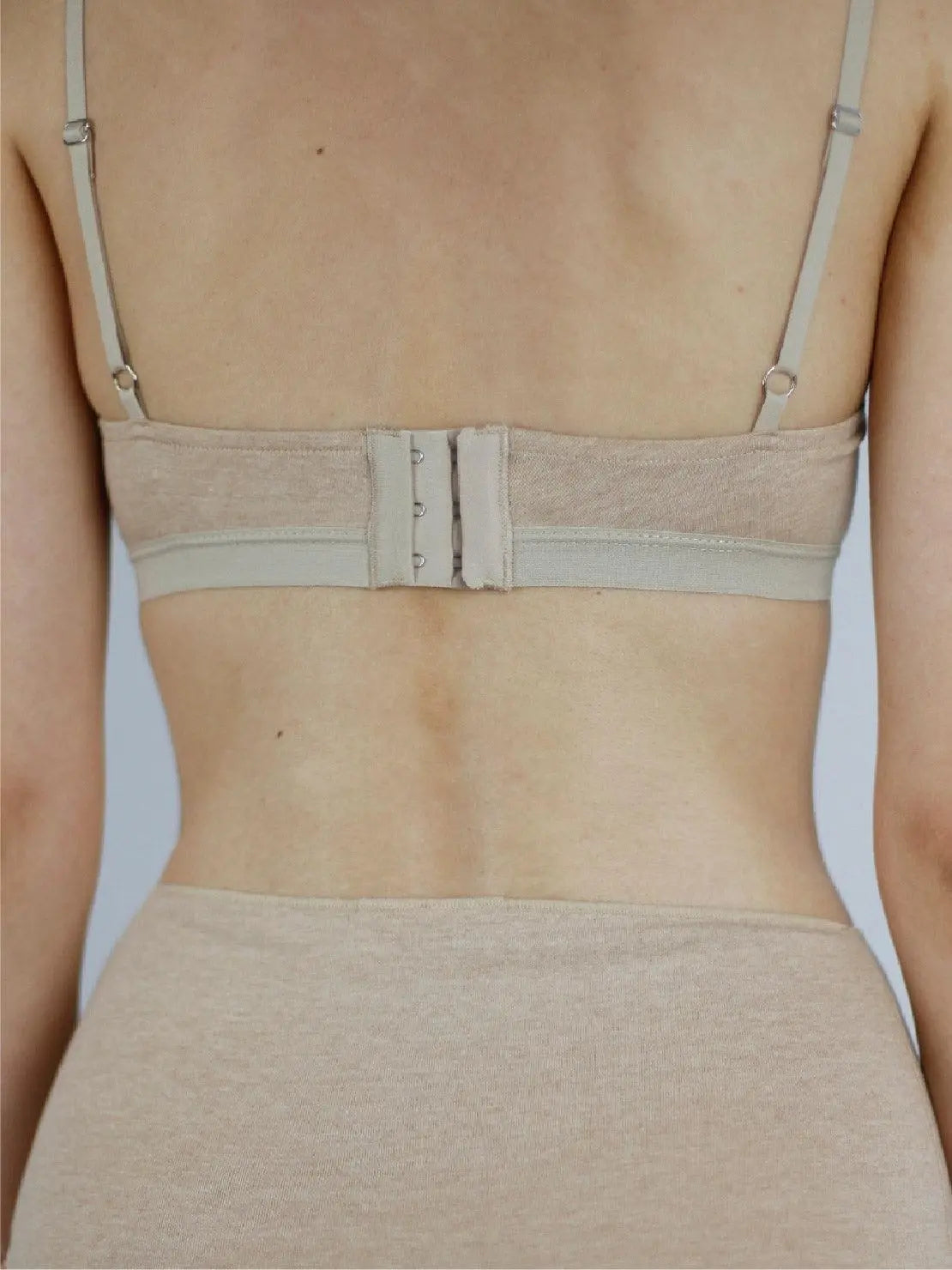 A person is shown from the shoulders to the midriff, wearing a light beige, strappy Earth Bandeau Organic Cotton Bra - Talk Under Light and high-waisted underwear. The plain gray background highlights the minimalist design of the soft, stretchable fabric. Discover this chic collection at Bassalstore in Barcelona.