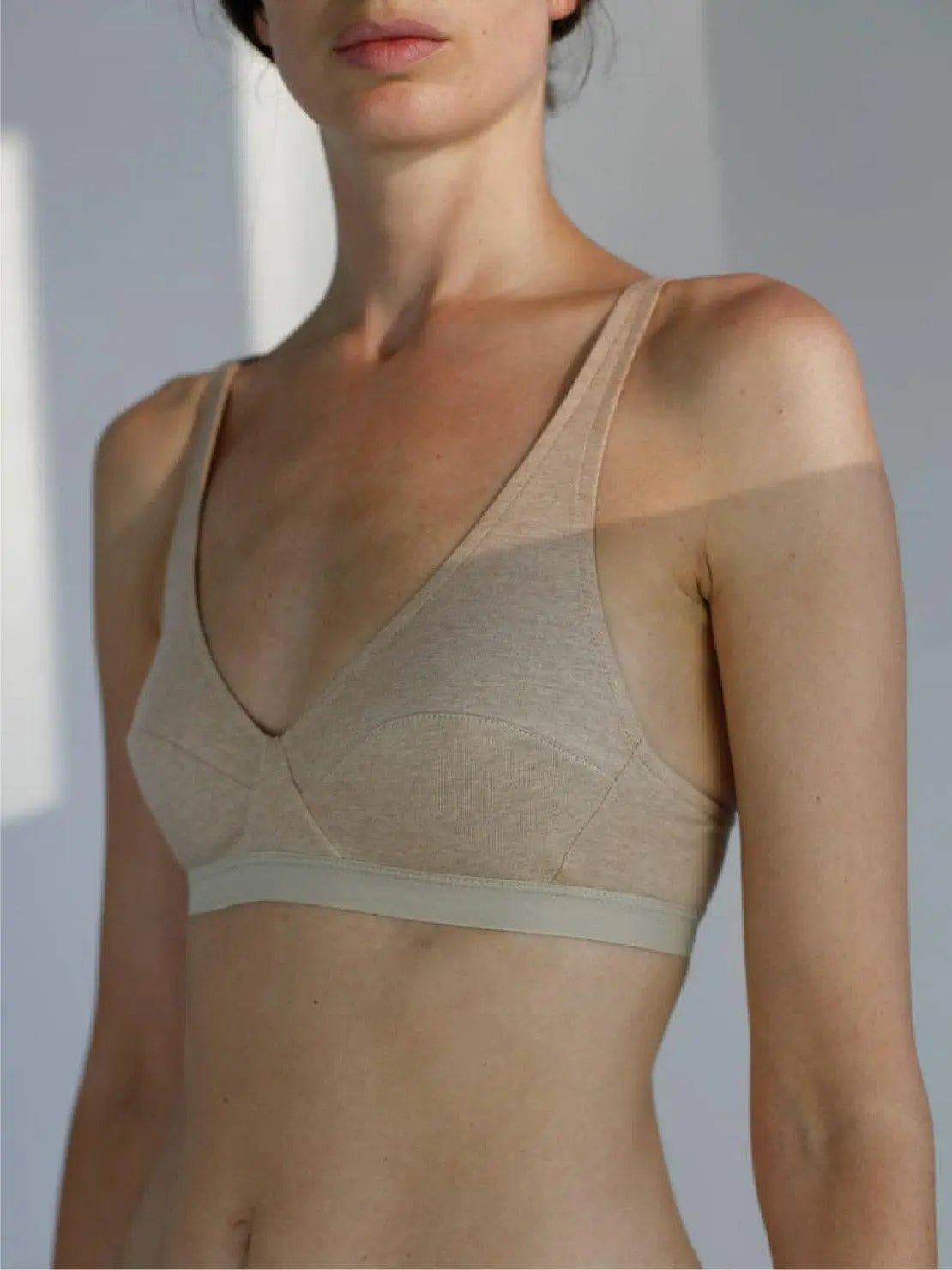 A person wearing an Earth Mia Organic Cotton Bra - Talk Under Light with a deep V-neckline and wide straps is seen from the shoulders up, possibly browsing in a Barcelona store. The person has a slender build and stands against a neutral background, while the lighting creates shadows and highlights on the skin and fabric.