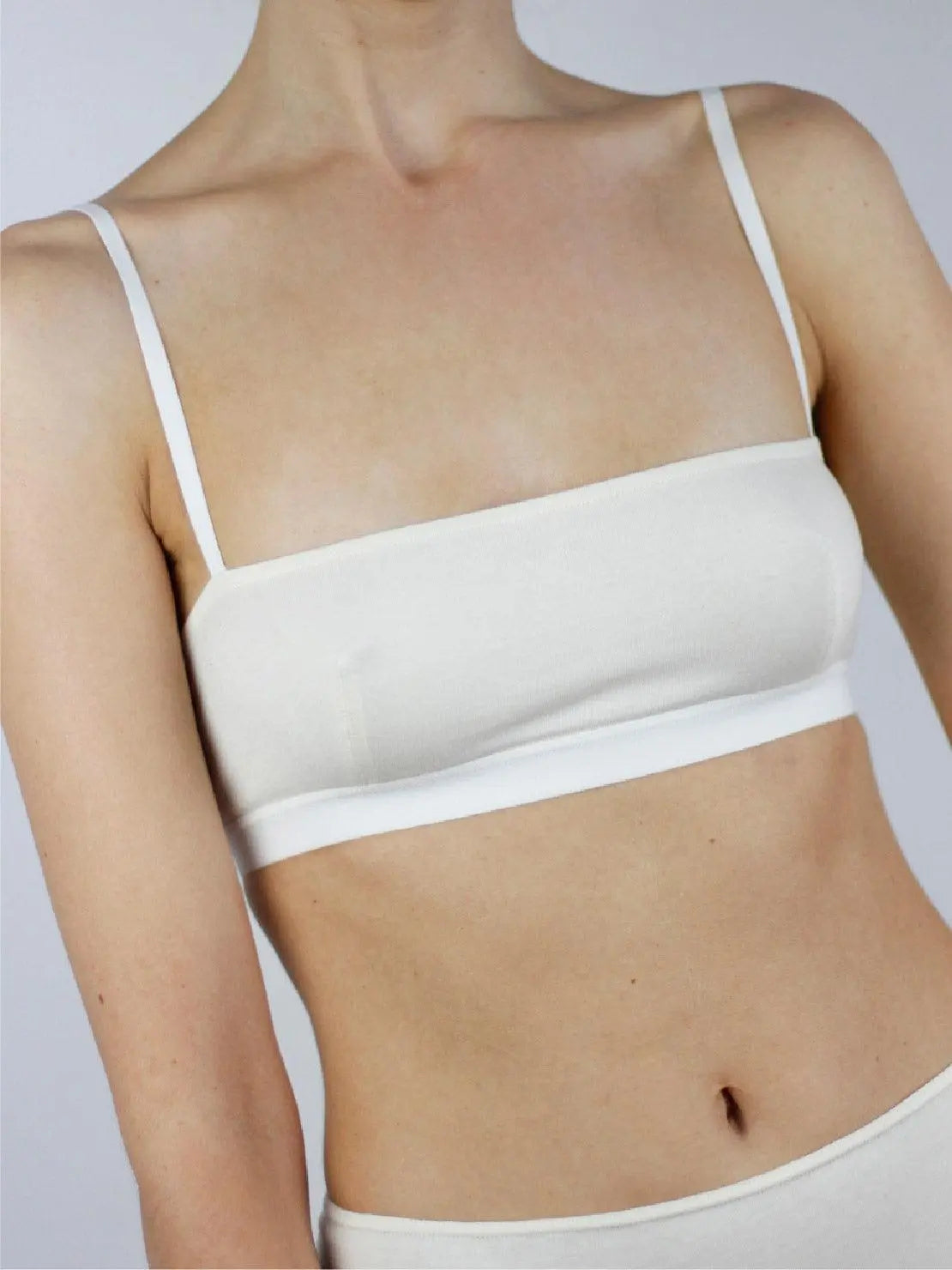 A person is wearing a white, minimalist Ecru Bandeau Organic Cotton Bra - Talk Under Light with thin straps. The image focuses on the upper torso, revealing the bra's simple and clean design. The background is a plain light grey, enhancing the focus on the Ecru Bandeau Organic Cotton Bra - Talk Under Light, available exclusively at BassalStore Barcelona.