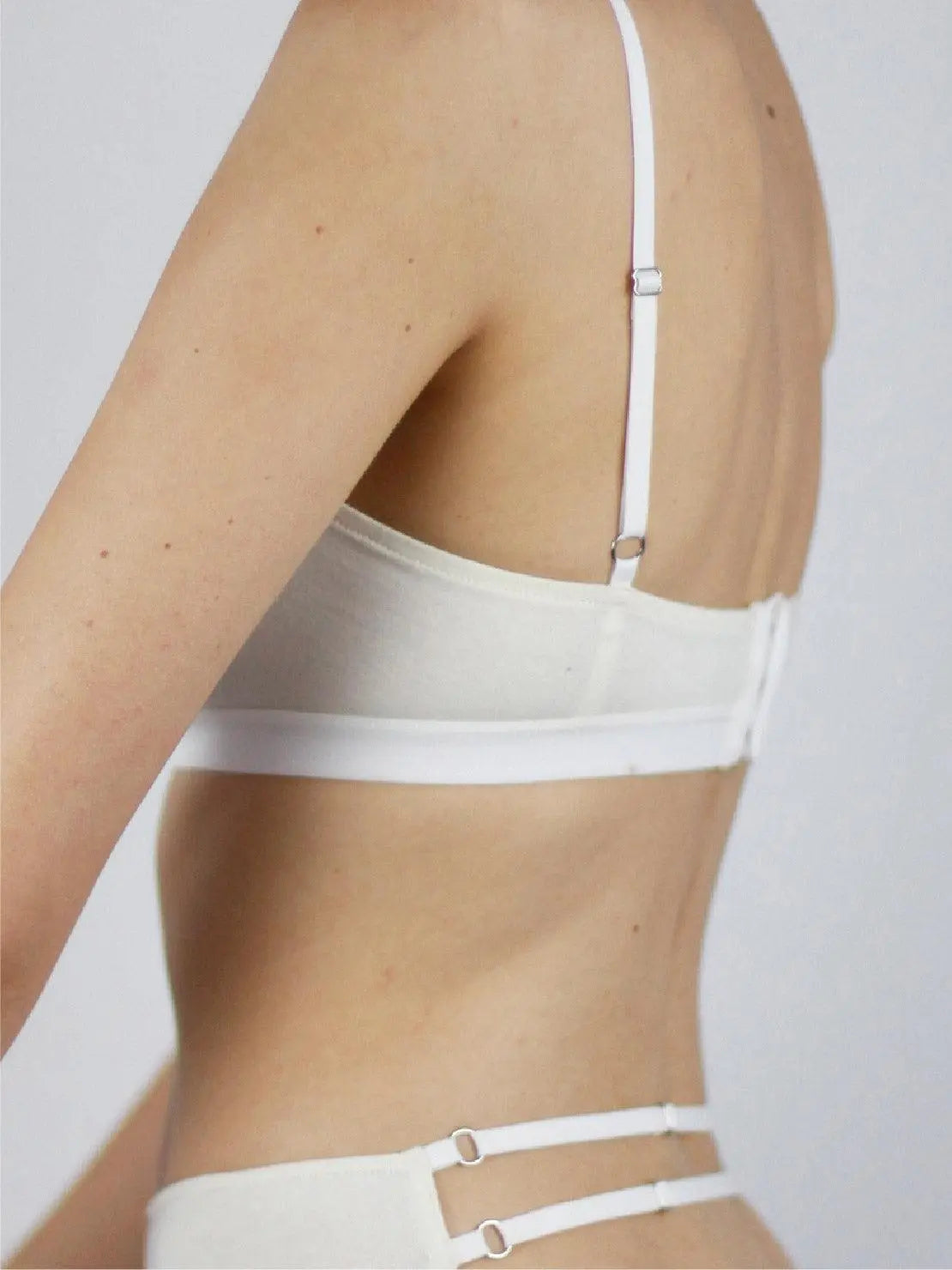 A person is wearing a white, minimalist Ecru Bandeau Organic Cotton Bra - Talk Under Light with thin straps. The image focuses on the upper torso, revealing the bra's simple and clean design. The background is a plain light grey, enhancing the focus on the Ecru Bandeau Organic Cotton Bra - Talk Under Light, available exclusively at BassalStore Barcelona.
