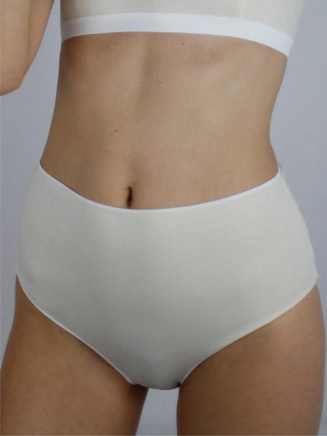 A person is shown from the torso down wearing Ecru Basic Highwaist Organic Cotton Briefs - Talk Under Light and a banded white bralette. The background is neutral, focusing attention on the minimalist undergarments available at Bassalstore in Barcelona.