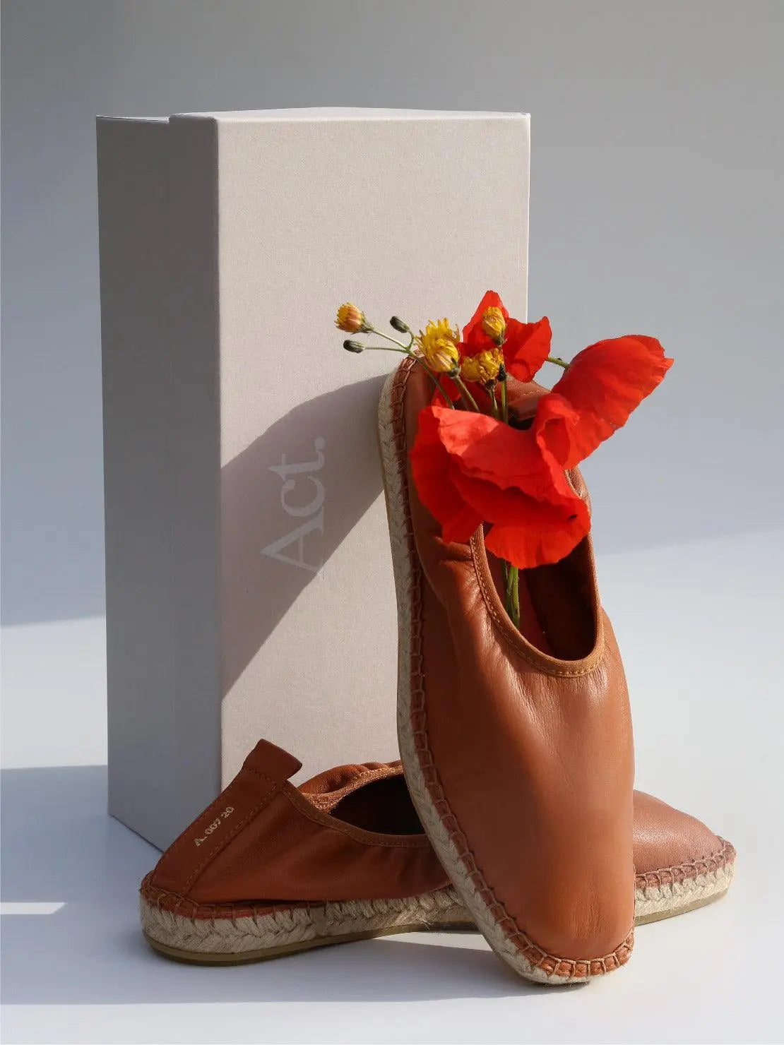 A pair of Edvard Brown Leather Espadrilles - Act Series with woven soles, one upright and the other resting on its side, next to a branded Act Series box. Red poppy flowers and yellow blossoms peek out from the upright shoe. Light background with soft shadows, perfect for your next visit to a chic Barcelona store like Bassalstore.