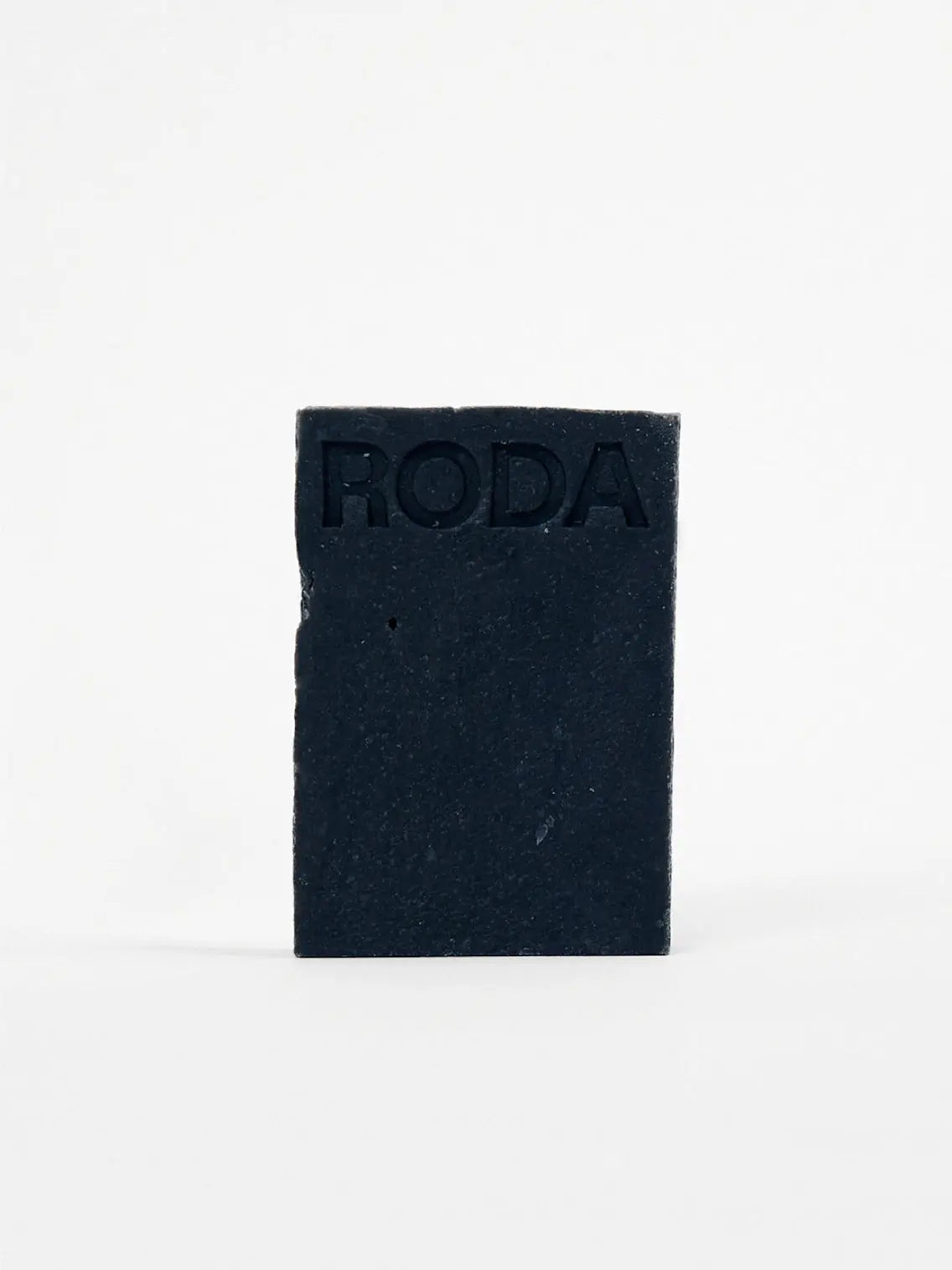 A rectangular, dark-colored bar of soap with the word "RODA" etched into the top section is standing upright against a plain white background, exuding an air of elegance reminiscent of a chic Barcelona boutique. This is the Face & Body Soap Bar - Activated Charcoal & Peppermint by Roda.