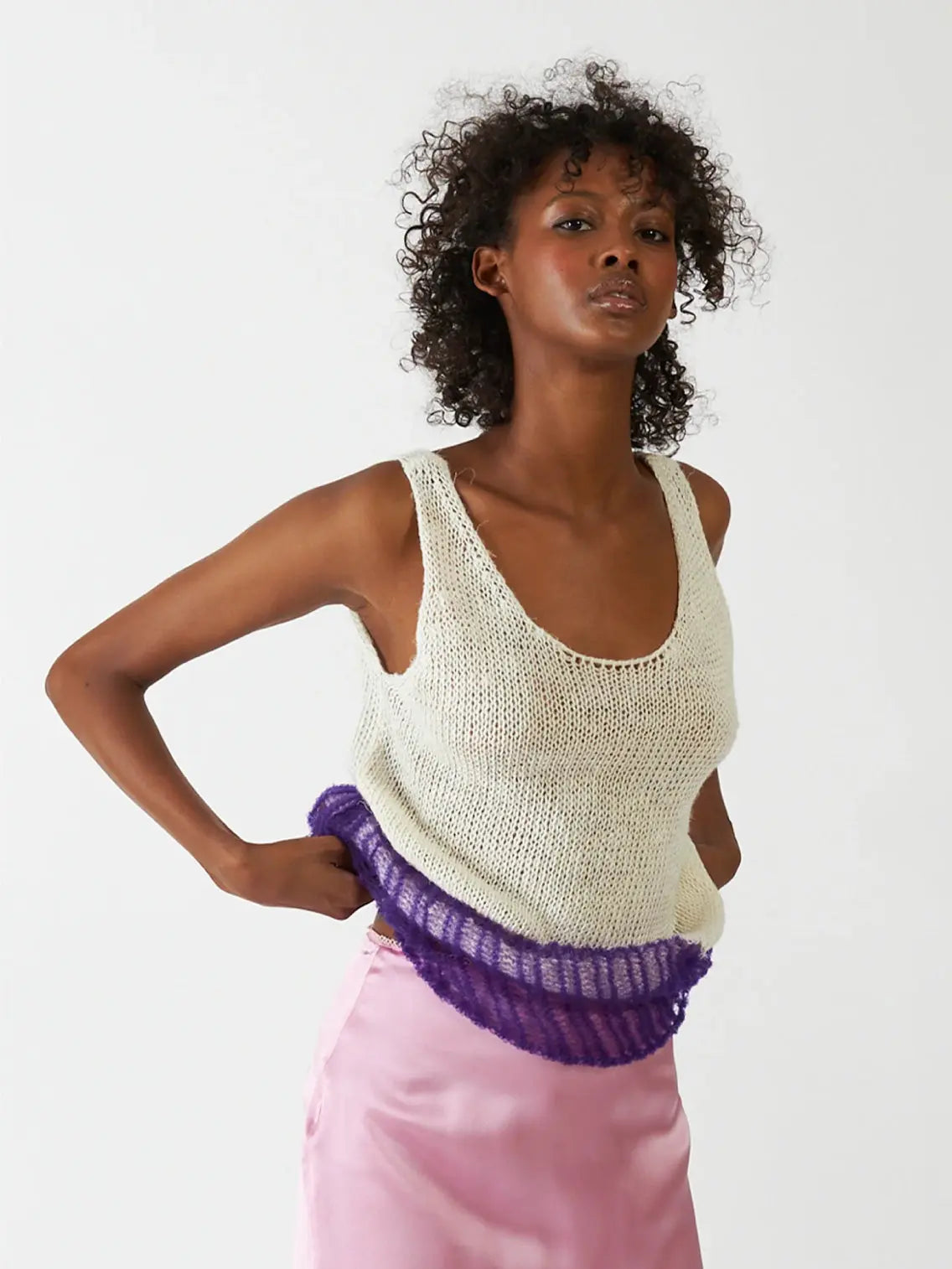 A person with curly hair is wearing a sleeveless, scoop-neck, off-white knit top with a purple ruffle at the bottom. They also have on a light pink satin skirt and are posing against a plain white background, showcasing chic fashion available at bassalstore in Barcelona. The featured top is the Flin Top Purple by Bielo.
