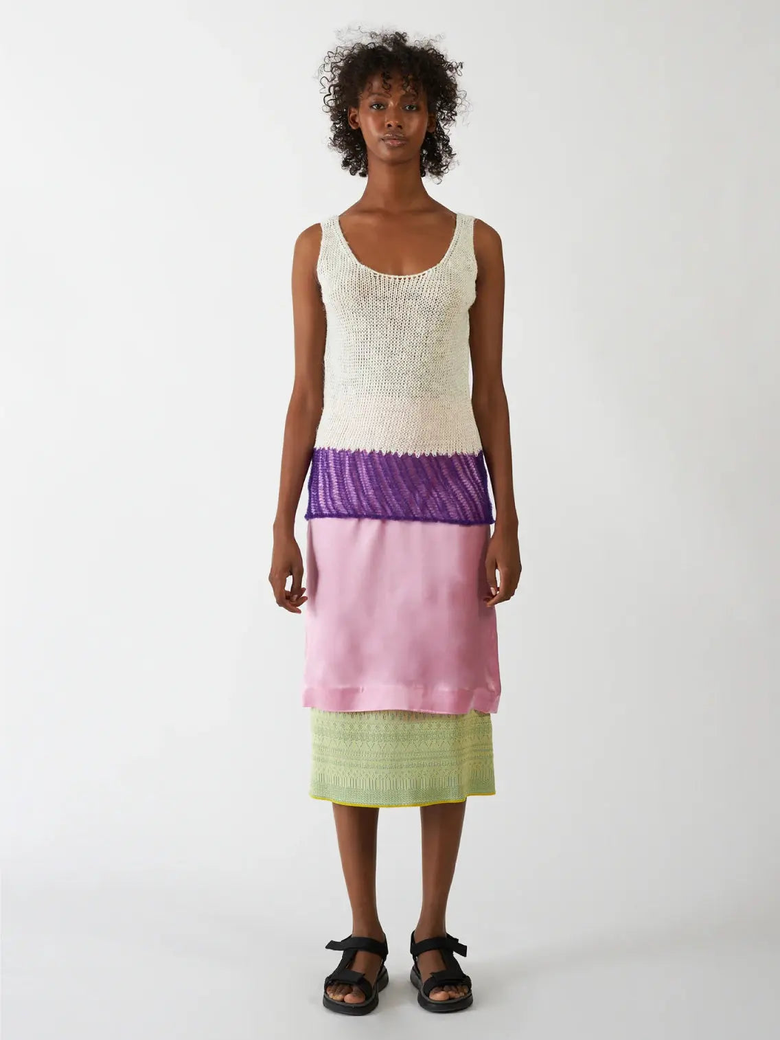 A person with curly hair is wearing a sleeveless, scoop-neck, off-white knit top with a purple ruffle at the bottom. They also have on a light pink satin skirt and are posing against a plain white background, showcasing chic fashion available at bassalstore in Barcelona. The featured top is the Flin Top Purple by Bielo.