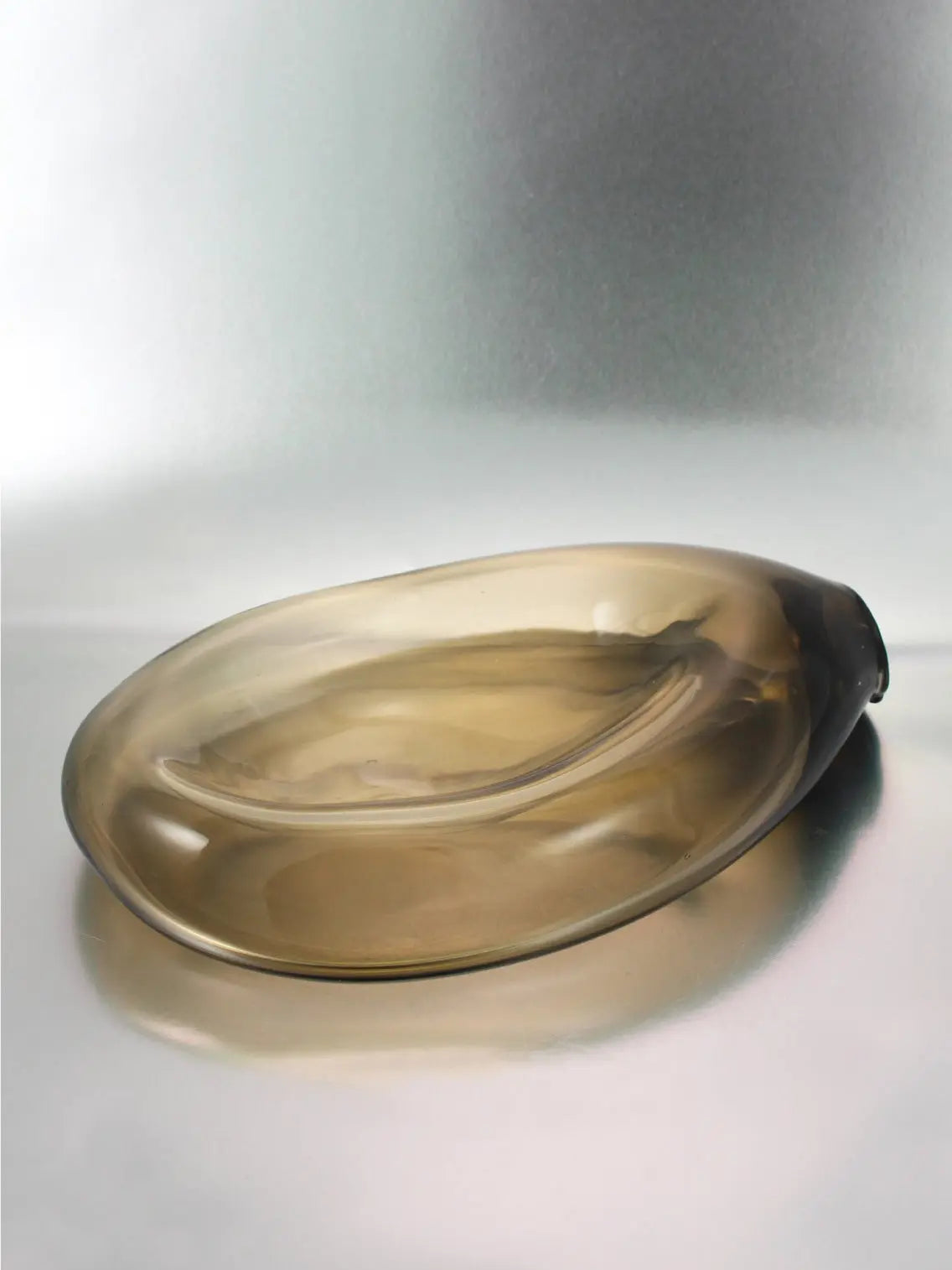 An elongated, translucent, brownish glass Gil Plate Olive by Nathalie Schreckenberg sits on a reflective surface. Available at Bassalstore in Barcelona, its shape is smooth and slightly asymmetrical, giving it a modern yet organic appearance. The gradient of the color intensifies toward one end of the plate.