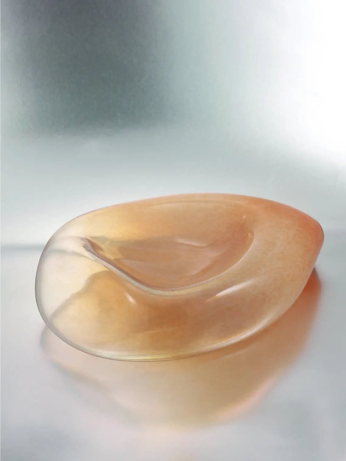 An abstract, smooth, and transparent glass sculpture from Nathalie Schreckenberg rests on a reflective surface. The Gil Plate Peach features a soft, organic shape with subtle gradients of amber hues blending into clear glass, creating a serene and fluid visual effect. Perfect for any Barcelona-inspired decor.