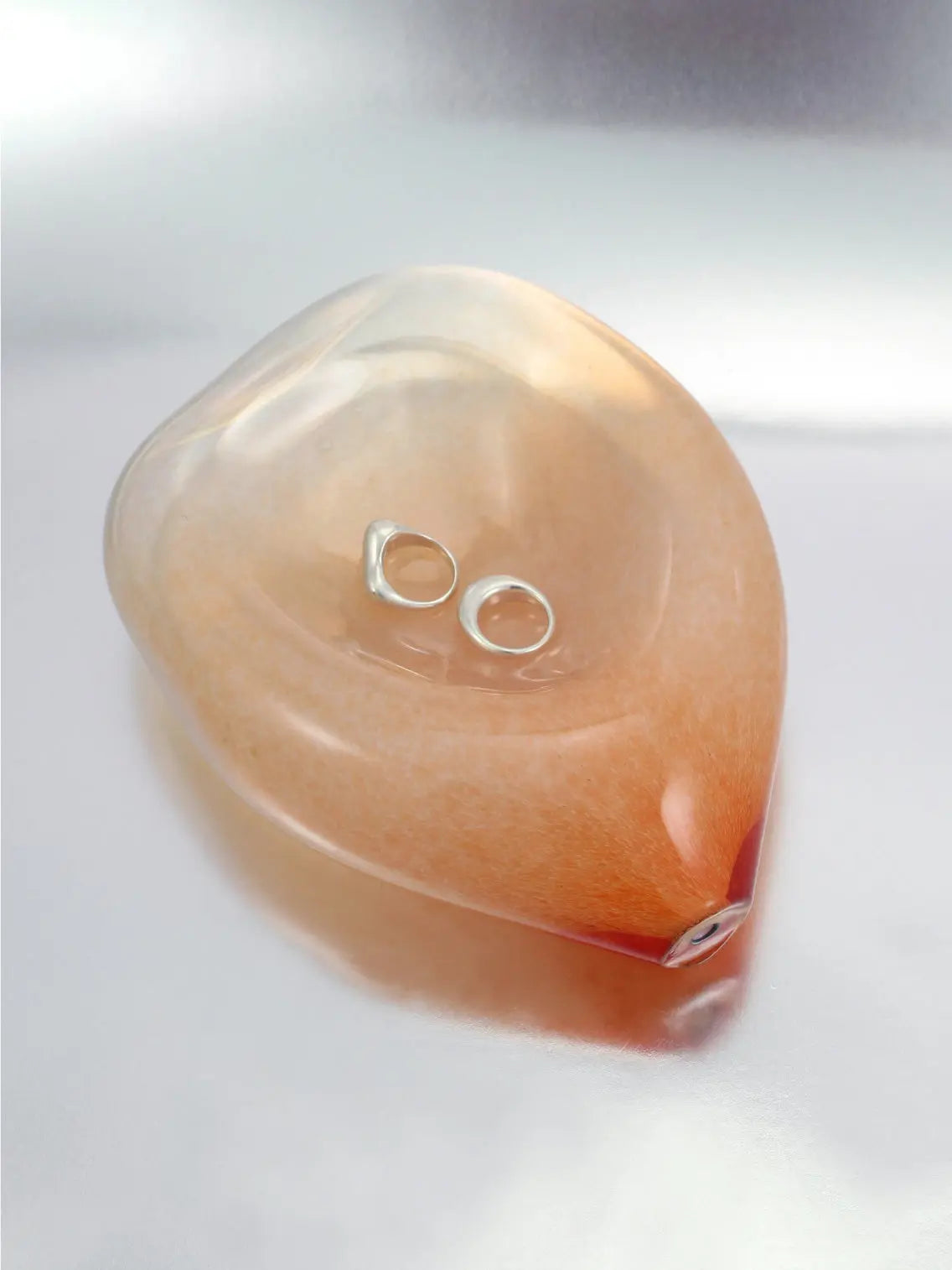 An abstract, smooth, and transparent glass sculpture from Nathalie Schreckenberg rests on a reflective surface. The Gil Plate Peach features a soft, organic shape with subtle gradients of amber hues blending into clear glass, creating a serene and fluid visual effect. Perfect for any Barcelona-inspired decor.