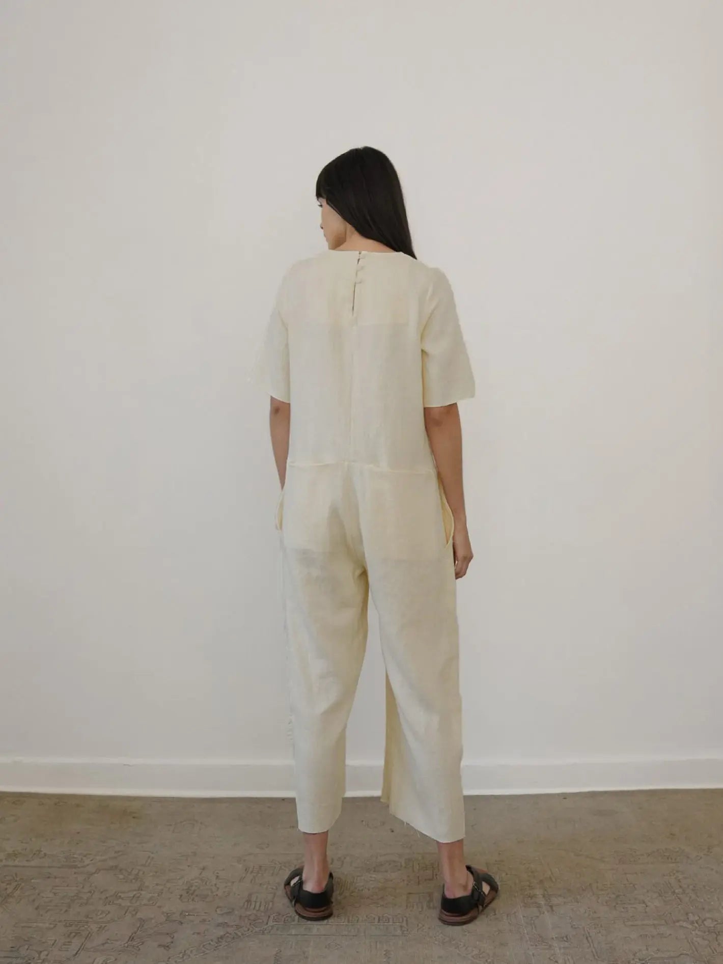 A woman with long dark hair stands facing a plain white wall, inside what appears to be a well-lit store in Barcelona. She is wearing the June Jumpsuit Beige by Zii Ropa and black sandals. The floor is light brown, possibly carpeted.