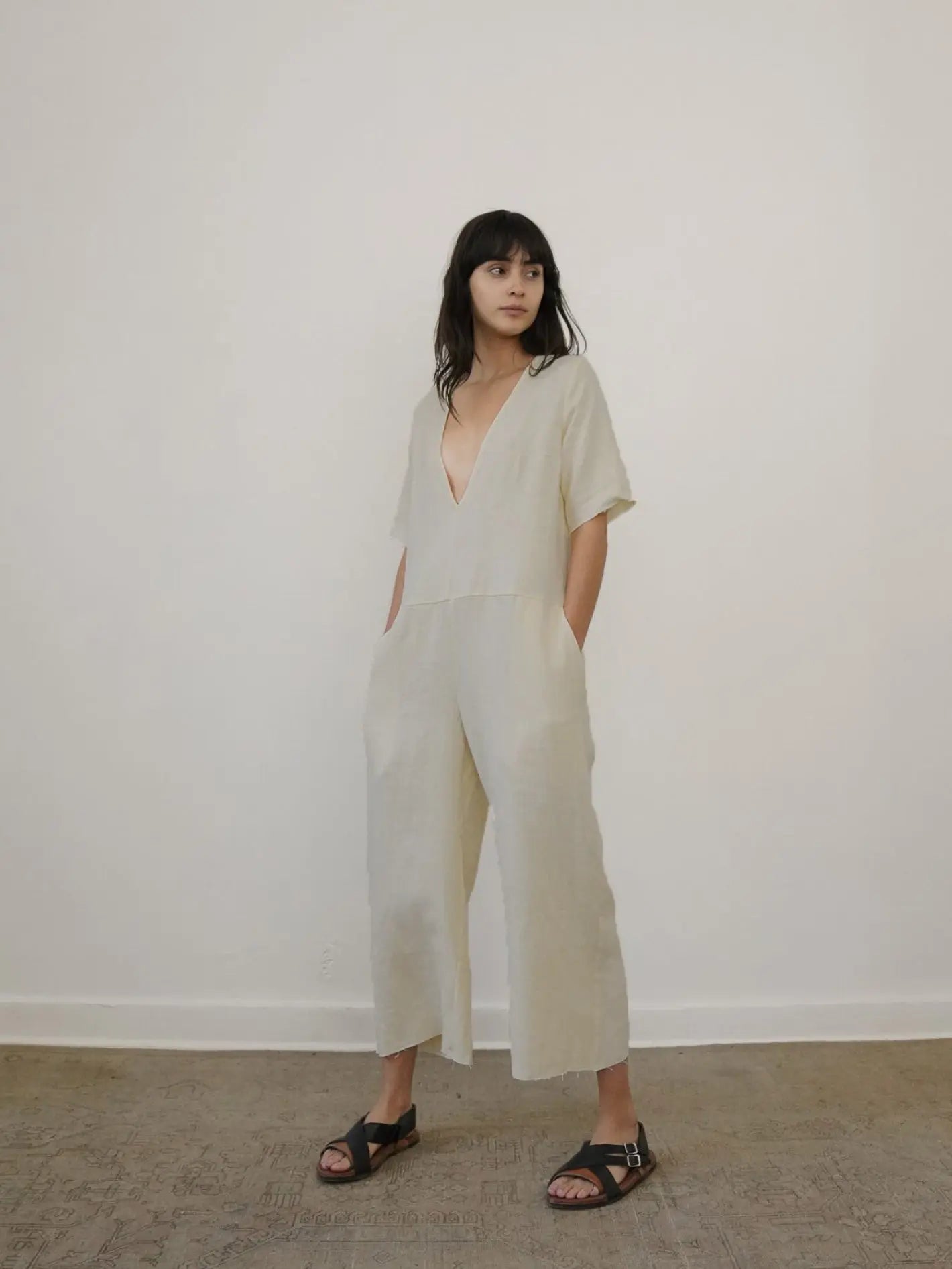 A woman with long dark hair stands facing a plain white wall, inside what appears to be a well-lit store in Barcelona. She is wearing the June Jumpsuit Beige by Zii Ropa and black sandals. The floor is light brown, possibly carpeted.