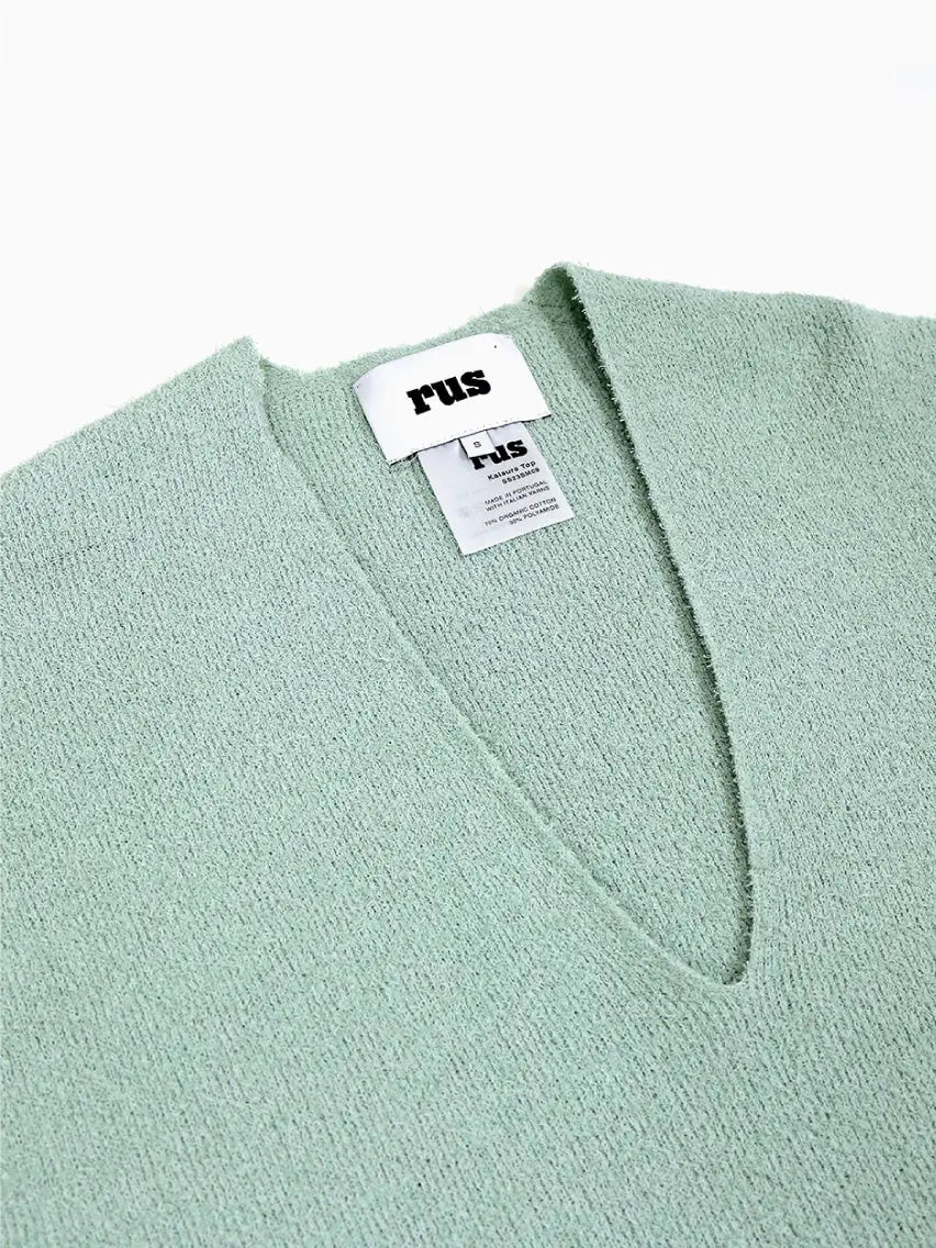 A Katsura Top Green with a deep V-neck and a fringe trim at the hem. The label inside the collar area reads "Rus," and additional garment care and size tags are visible underneath. This chic piece, available at BassalStore in Barcelona, is laid flat against a plain white background.