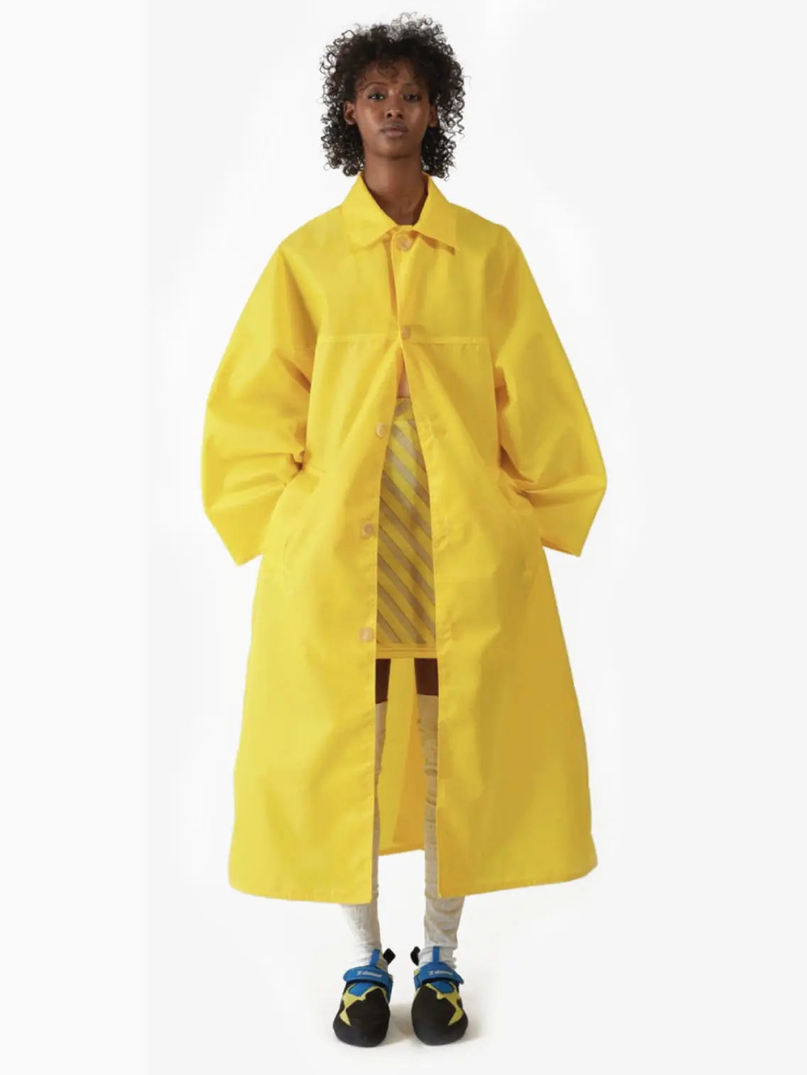 A person with short, curly hair is wearing a long, oversized Kim Long Coat Yellow by Bielo that reaches below the knees. They are standing against a plain white background, with hands in the coat pockets, revealing a hint of a striped outfit underneath—perfectly suited for strolling through Barcelona or visiting Bassalstore.