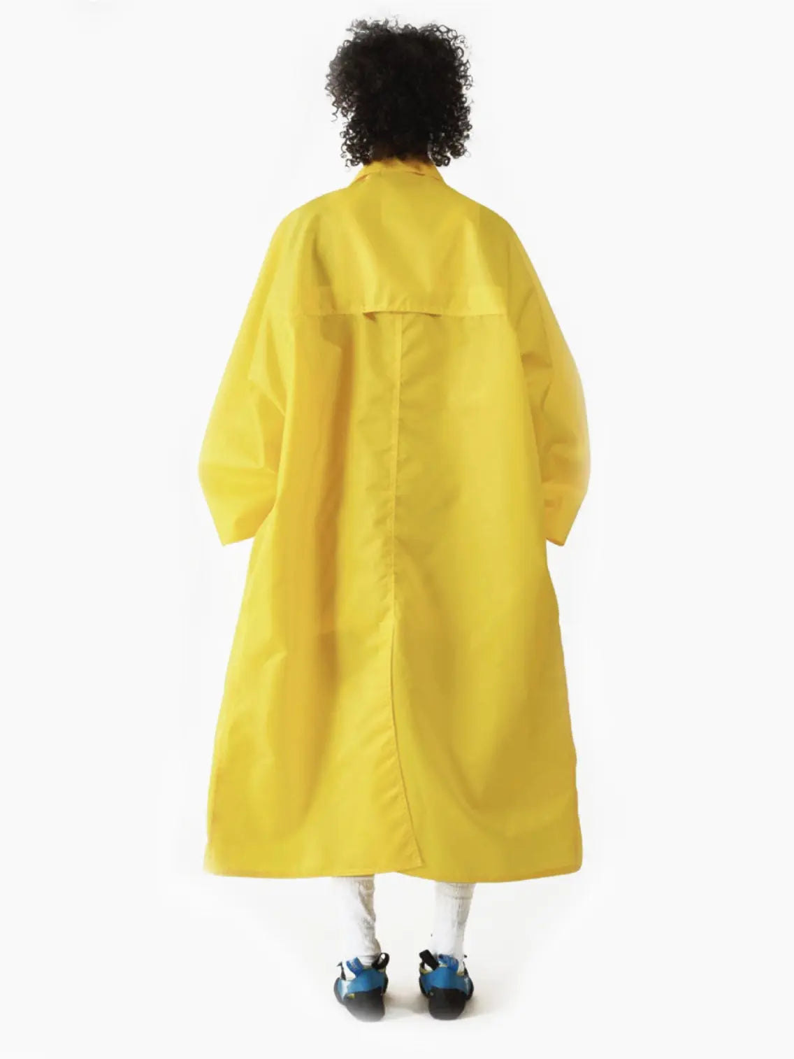 A person with short, curly hair is wearing a long, oversized Kim Long Coat Yellow by Bielo that reaches below the knees. They are standing against a plain white background, with hands in the coat pockets, revealing a hint of a striped outfit underneath—perfectly suited for strolling through Barcelona or visiting Bassalstore.