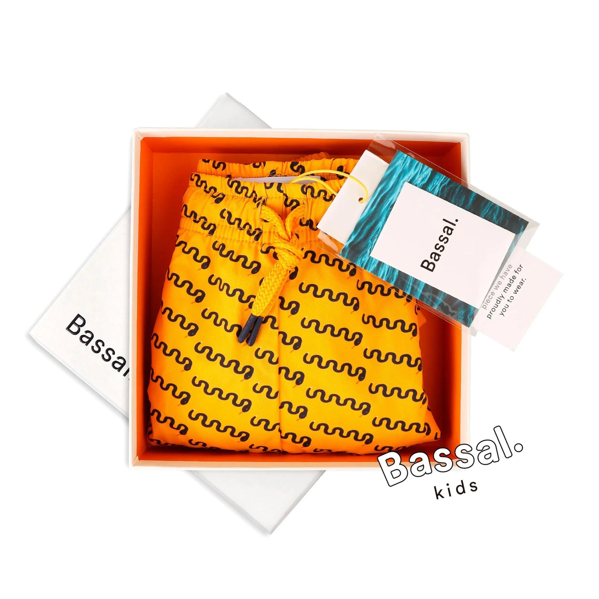An image of a pair of bright yellow, patterned shorts with black wavy lines, neatly packaged in an orange box. The box sits on a white surface next to a white envelope with the text "Bassal. kids" visible. A tag with the brand name "Bassal." is attached to the Mamba Kids Swimwear, spotlighting their availability at bassalstore in Barcelona.