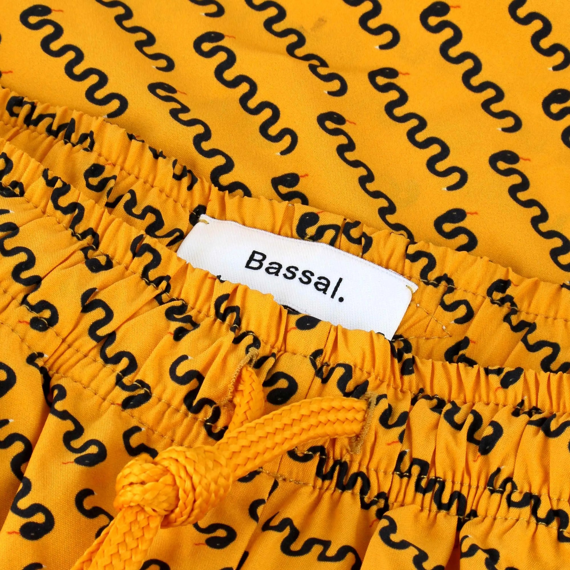 An open white box from bassalstore reveals a folded yellow Mamba Swimwear patterned with dark squiggly lines. The Mamba Swimwear has a blue drawstring and two tags: one reads "Bassal." and the other says, "proudly made for you to wear." The box lid proudly displays the brand name "Bassal" from Barcelona.