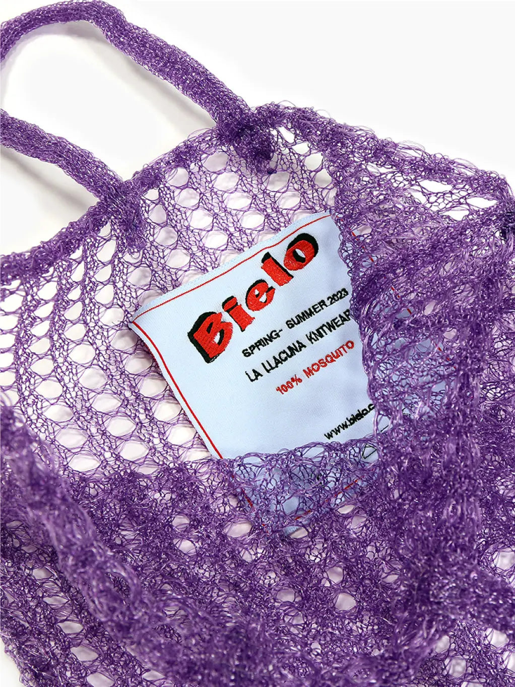 A purple, open-knit mesh market bag with two handles. The front features a white tag with red and black text, giving it a chic touch. This lightweight and stretchy bag is perfect for carrying groceries or other items—ideal for your next trip to the Barcelona store. Introducing the Bielo Mesh Tote Bag Lilac!