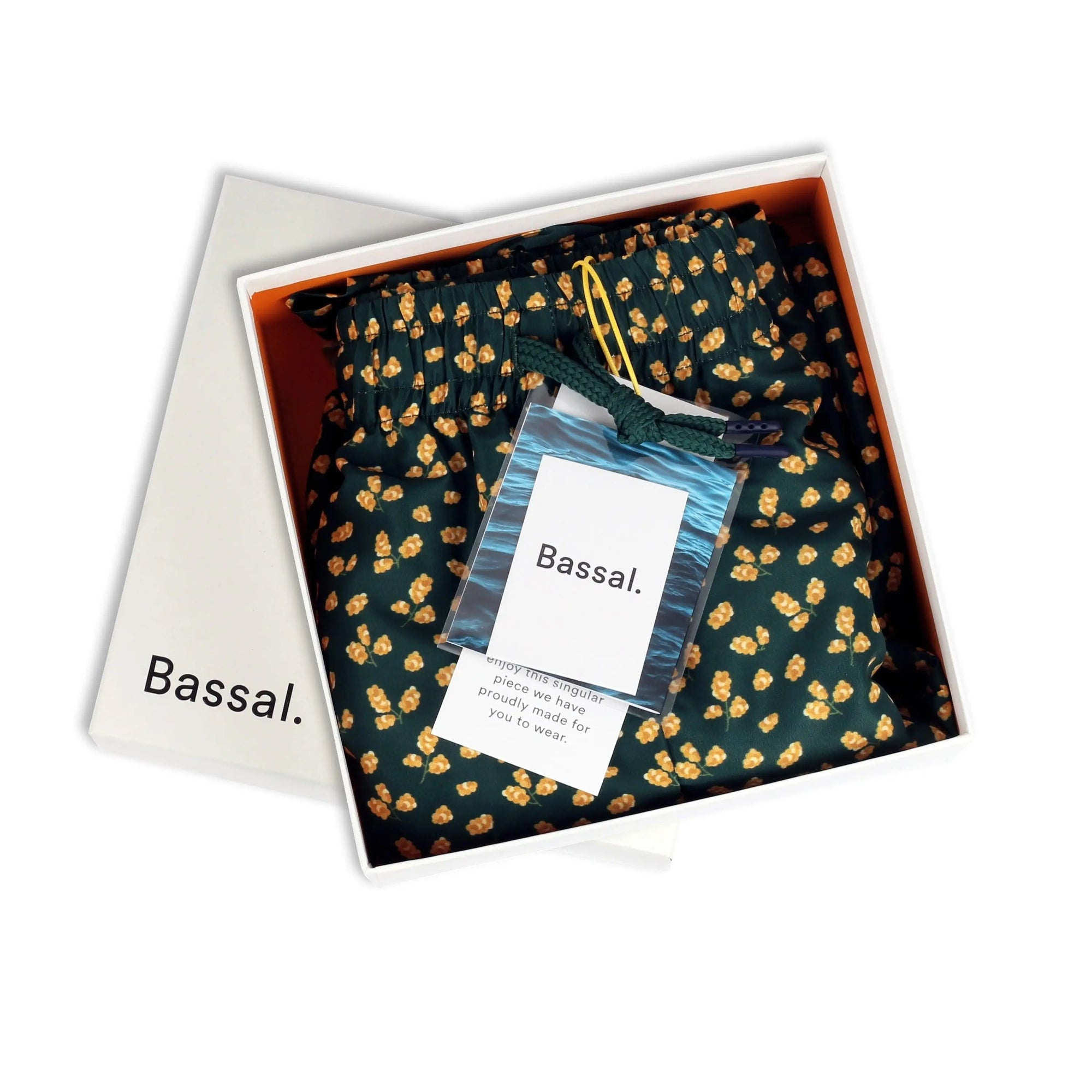 An open white box labeled "Bassal" reveals neatly folded dark green Mimosa Swimwear adorned with a yellow floral pattern. A smaller card with text lies on the swimwear, along with a small zipped bag containing a blue item. Discover this and more unique pieces at bassalstore, your go-to shop in Barcelona.