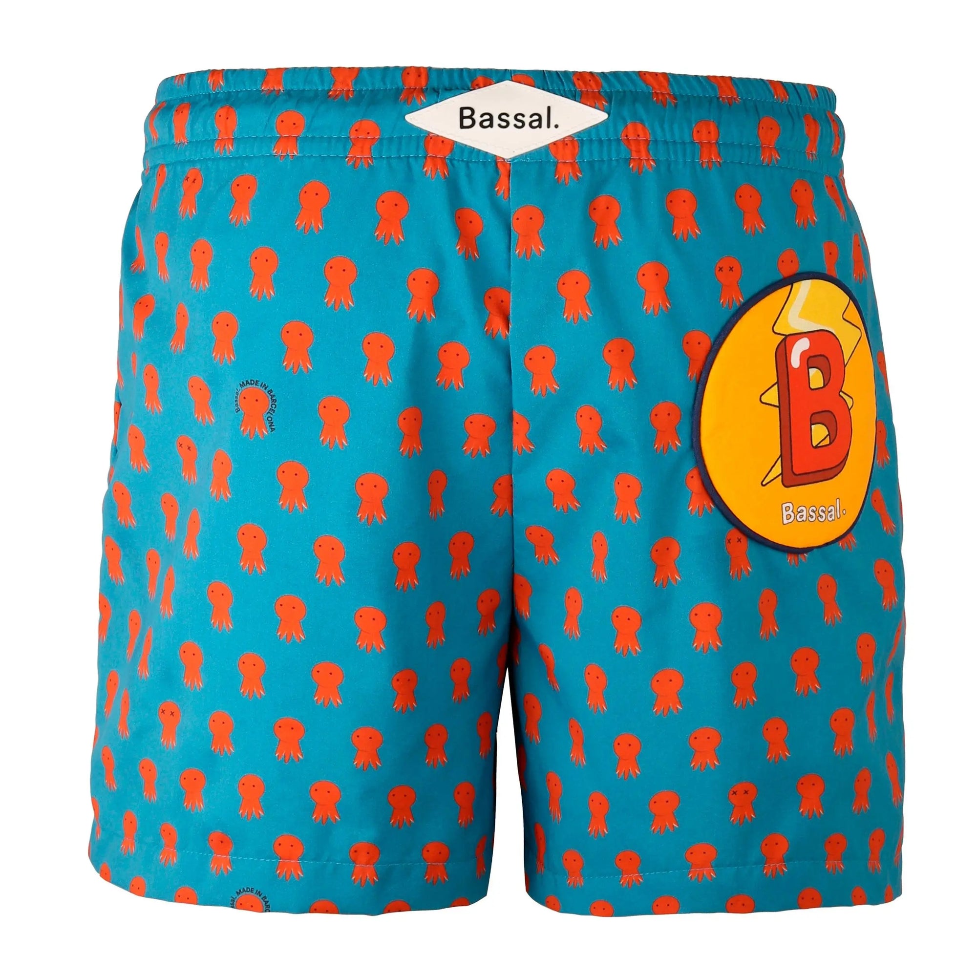 A pair of blue swim shorts with orange octopus prints is neatly folded inside a white box with an orange interior. The box, labeled "Bassal," hints at its origins from the chic Bassalstore in Barcelona. The Octo Kids Swimwear has a blue drawstring, and a note on top of the box appears to include text.