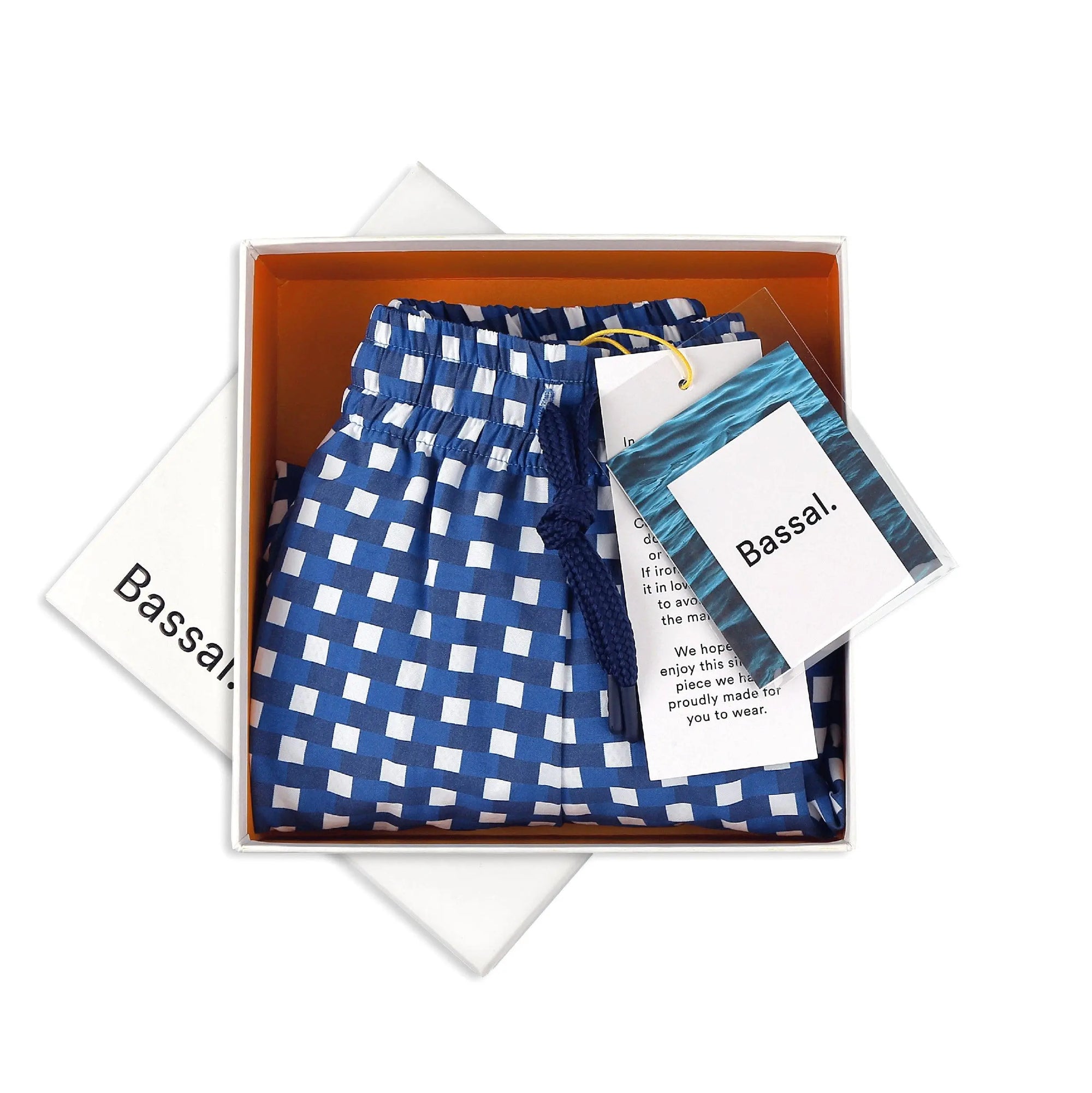 A pair of Paris Blue Swimwear is neatly folded inside an orange box with white edges. Attached to the swimwear are a white tag with the brand name "Bassal." and a card. The box lid, partially visible, also bears the brand name "Bassalstore," likely from their Barcelona store.